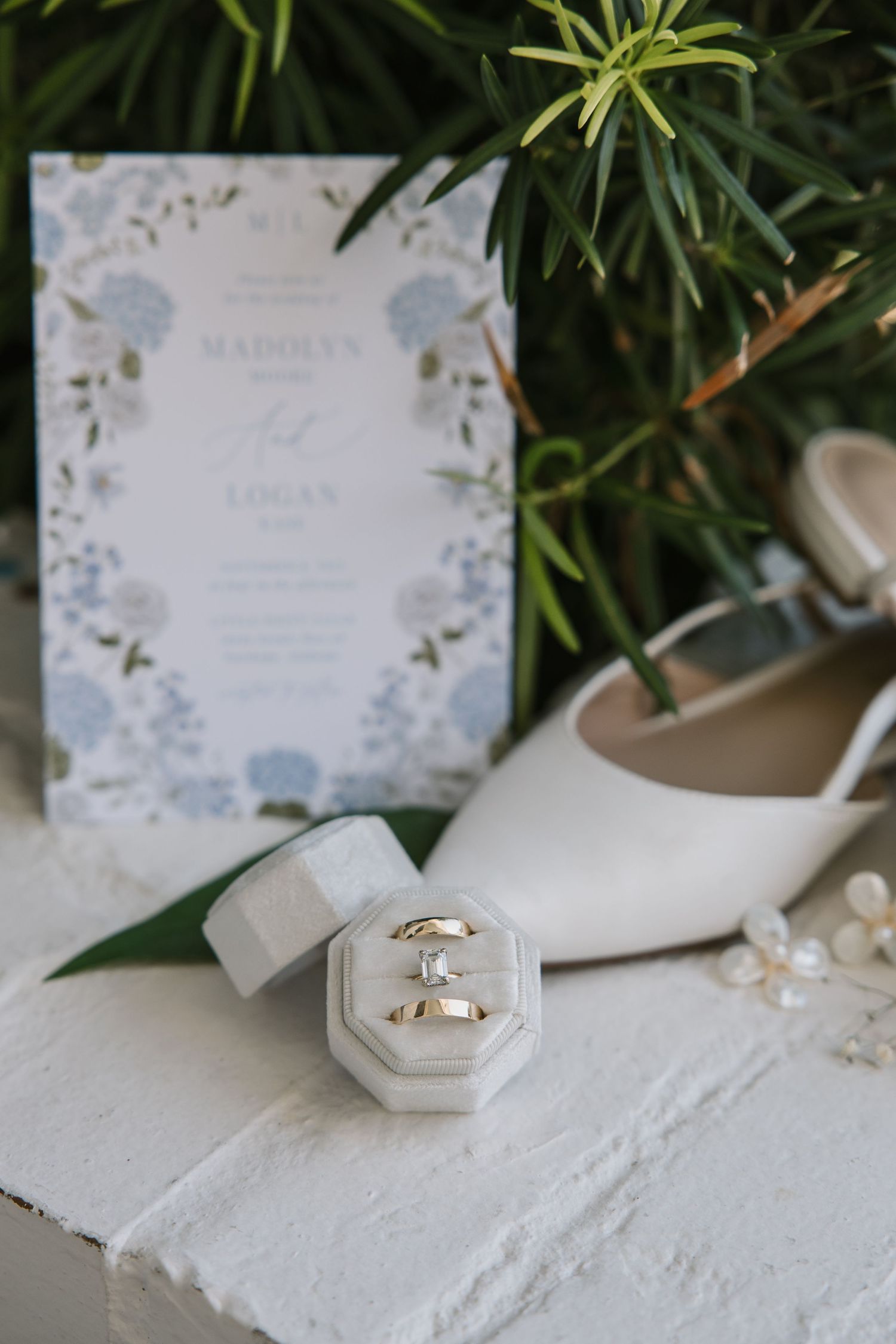 Elegant white and gold wedding rings displayed on textured surface with floral invitations and greenery in background.