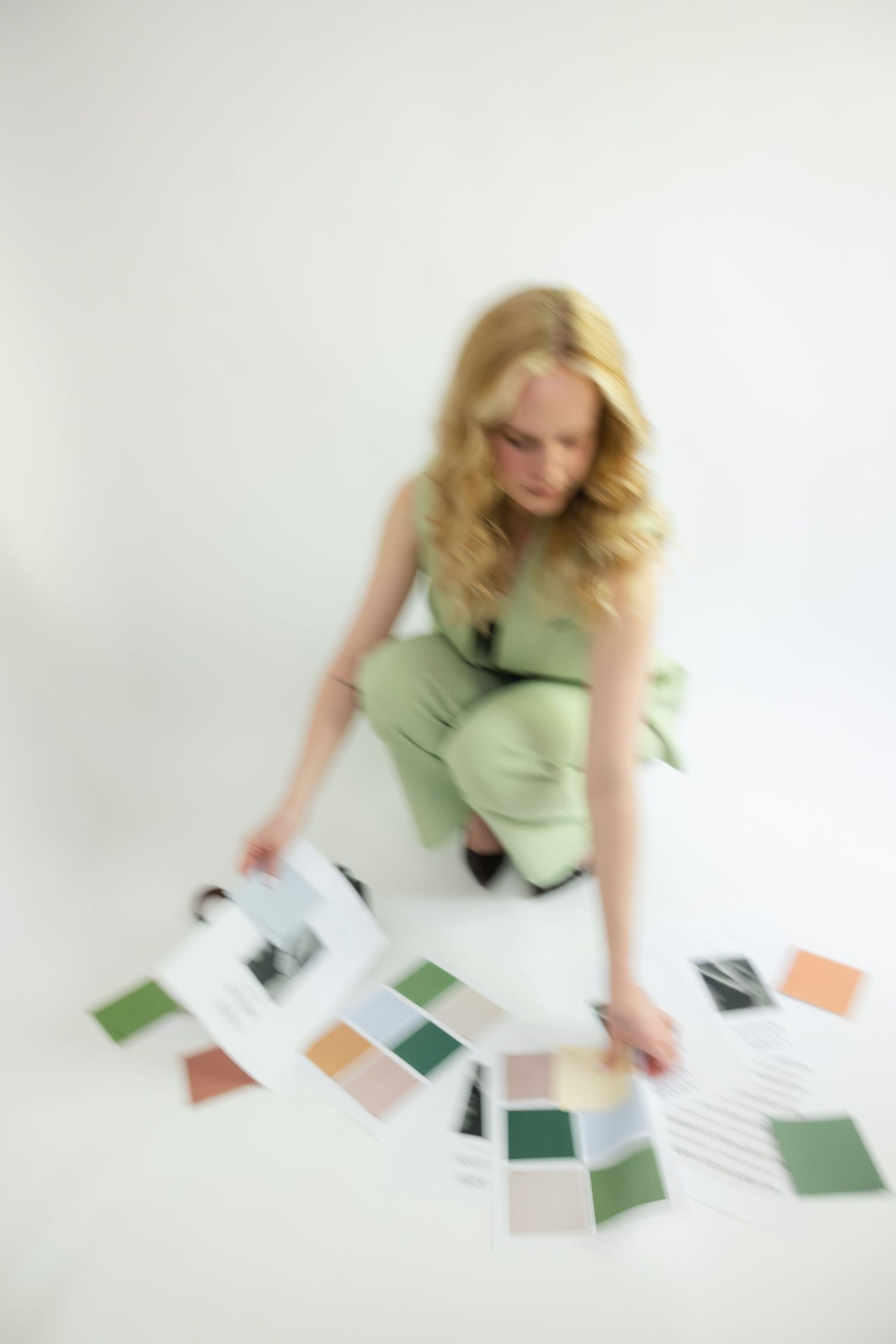 Person in mint green dress sits on white floor arranging scattered color swatches and papers in blurred motion shot.