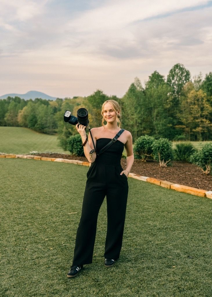 Professional photographer stands on a grassy hillside with camera equipment against a mountain backdrop in a black outfit.