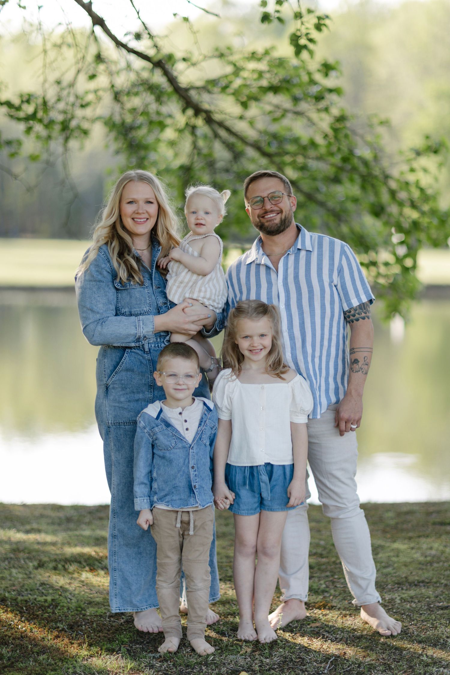 Family stands together by a serene lake on a sunny day, wearing coordinated blue and white outfits.