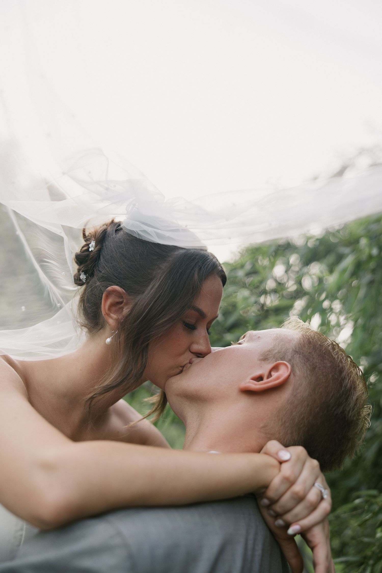 Romantic wedding couple sharing intimate kisses in a natural outdoor setting with flowing veil and greenery.