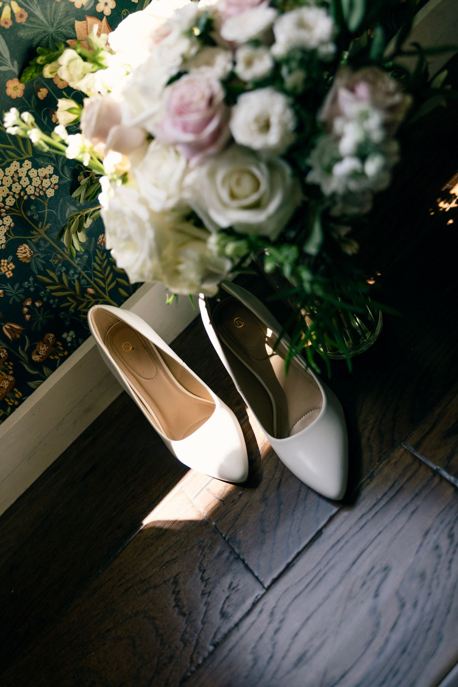 Elegant white bridal heels sit beside a fresh bouquet of pale roses and flowers on a dark wooden floor.