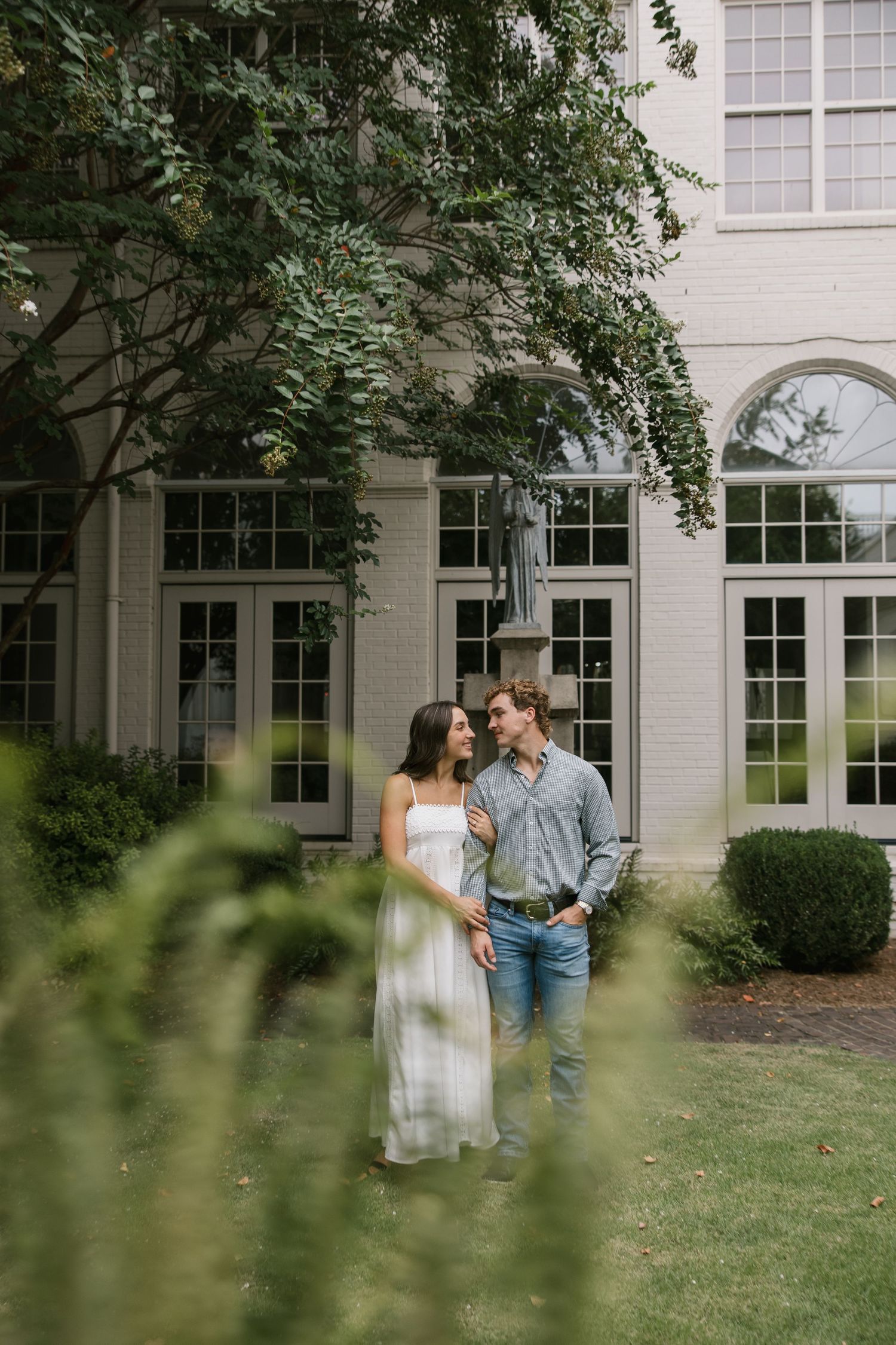 A couple shares an intimate moment in front of a white building with large windows surrounded by greenery.