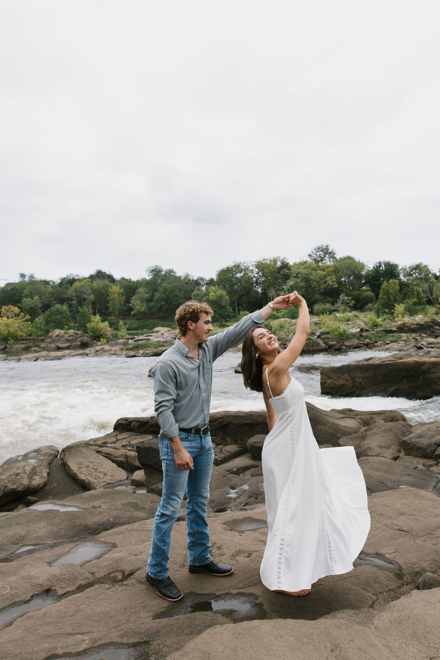 A couple dances joyfully together on rocky terrain near a river with trees in the background.