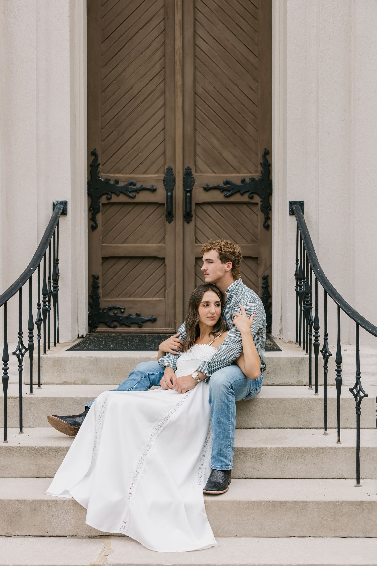 A happy couple sitting on church steps in wedding attire with white dress and denim jacket while leaning against pillars.