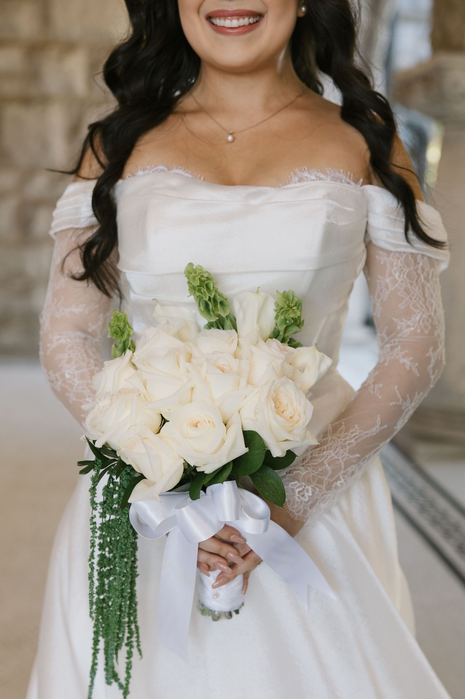 A bride in an off-shoulder lace wedding dress holds a stunning white rose bouquet with cascading greenery.