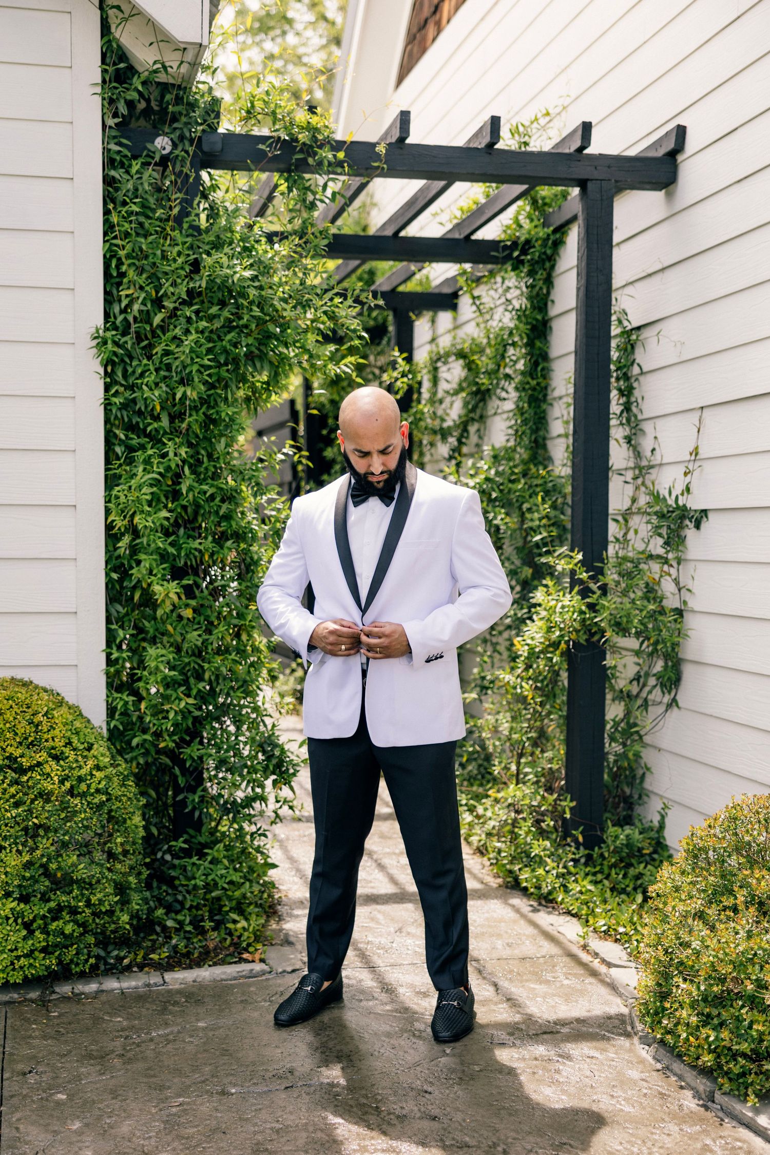 A person in a white tuxedo jacket and bow tie stands in a garden path beneath a dark pergola framed by greenery.