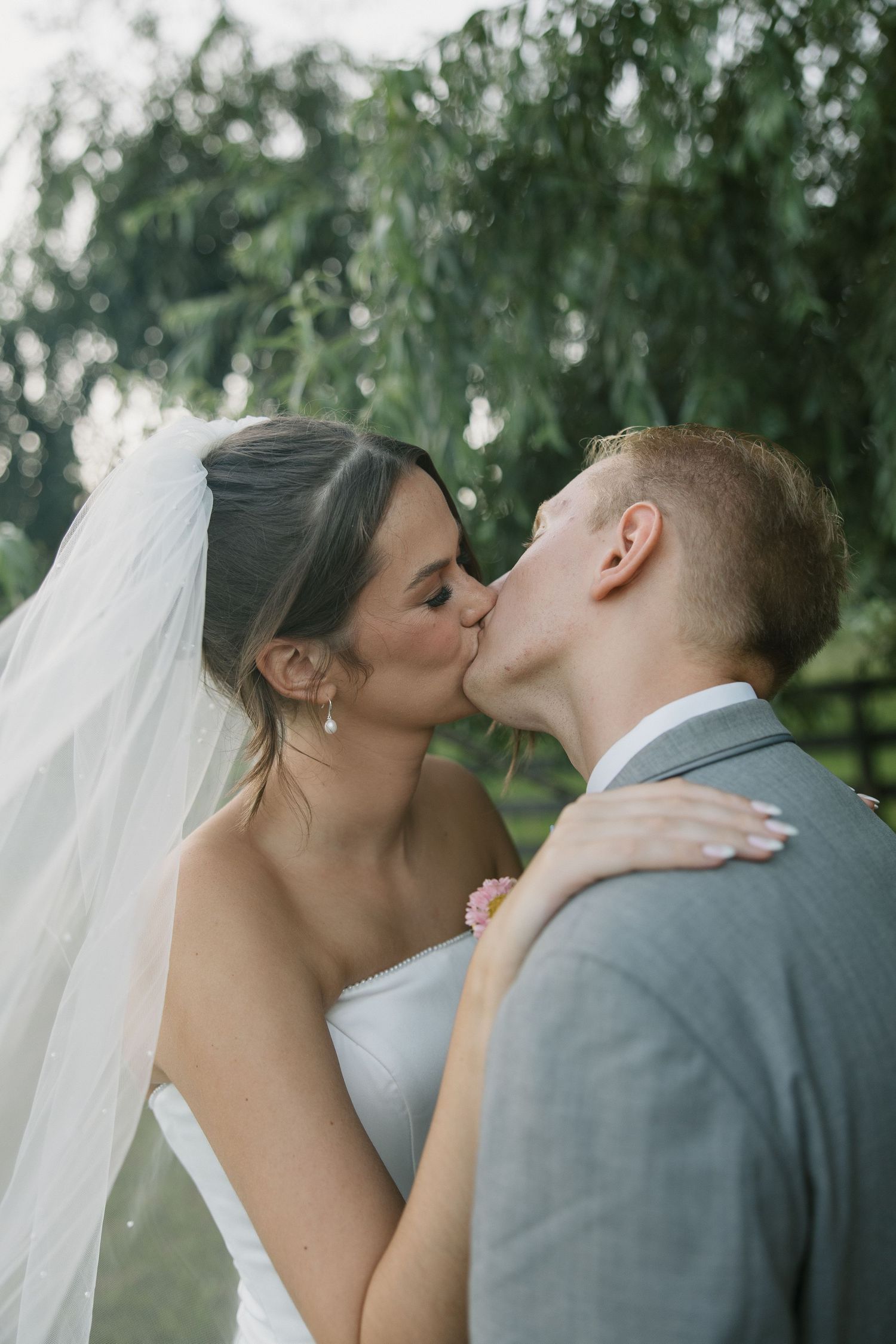 A tender kiss between newlyweds with a flowing white veil and grey suit against a soft green outdoor backdrop.
