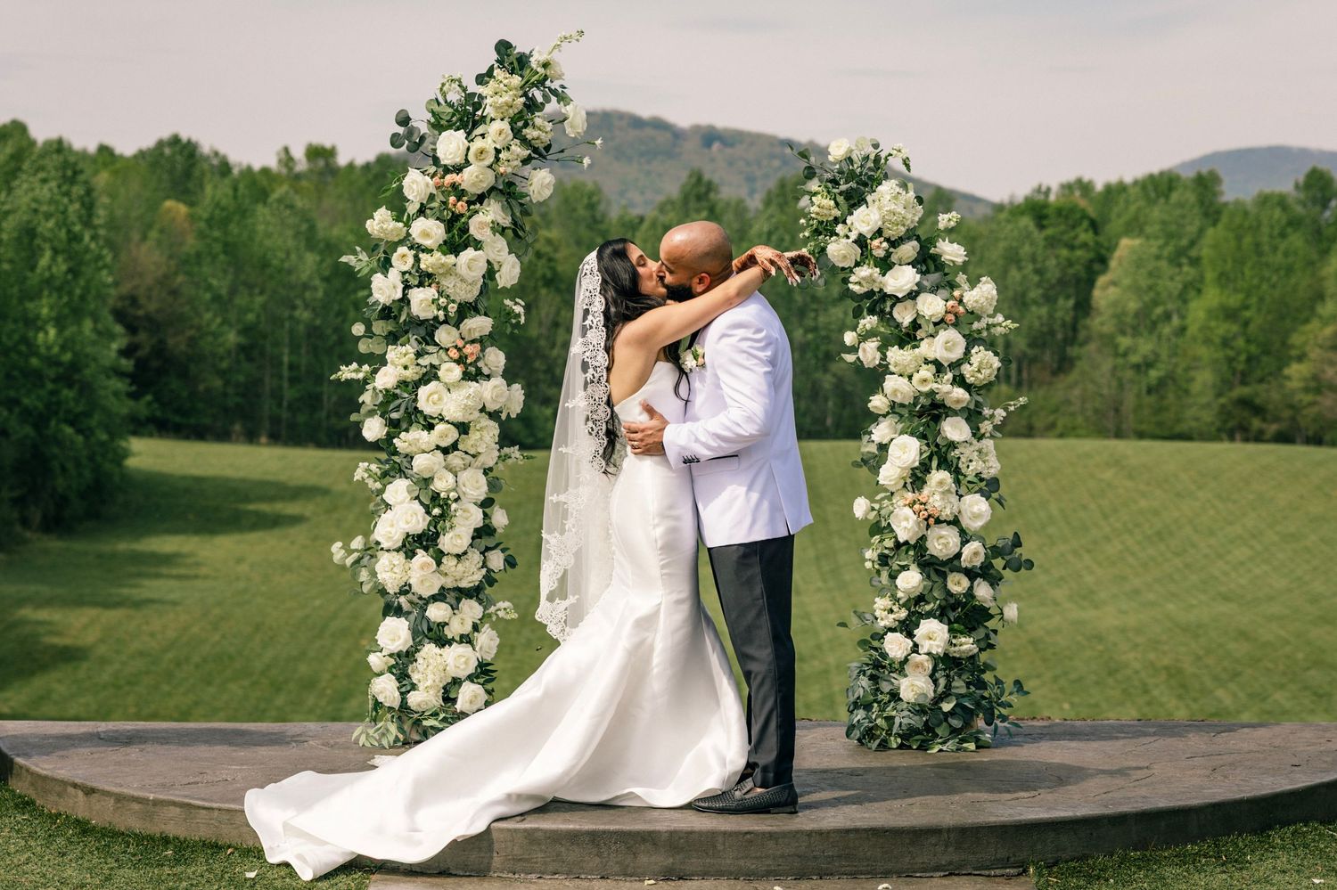 A romantic wedding ceremony at an outdoor venue with beautiful white floral arches and mountain views in the background.