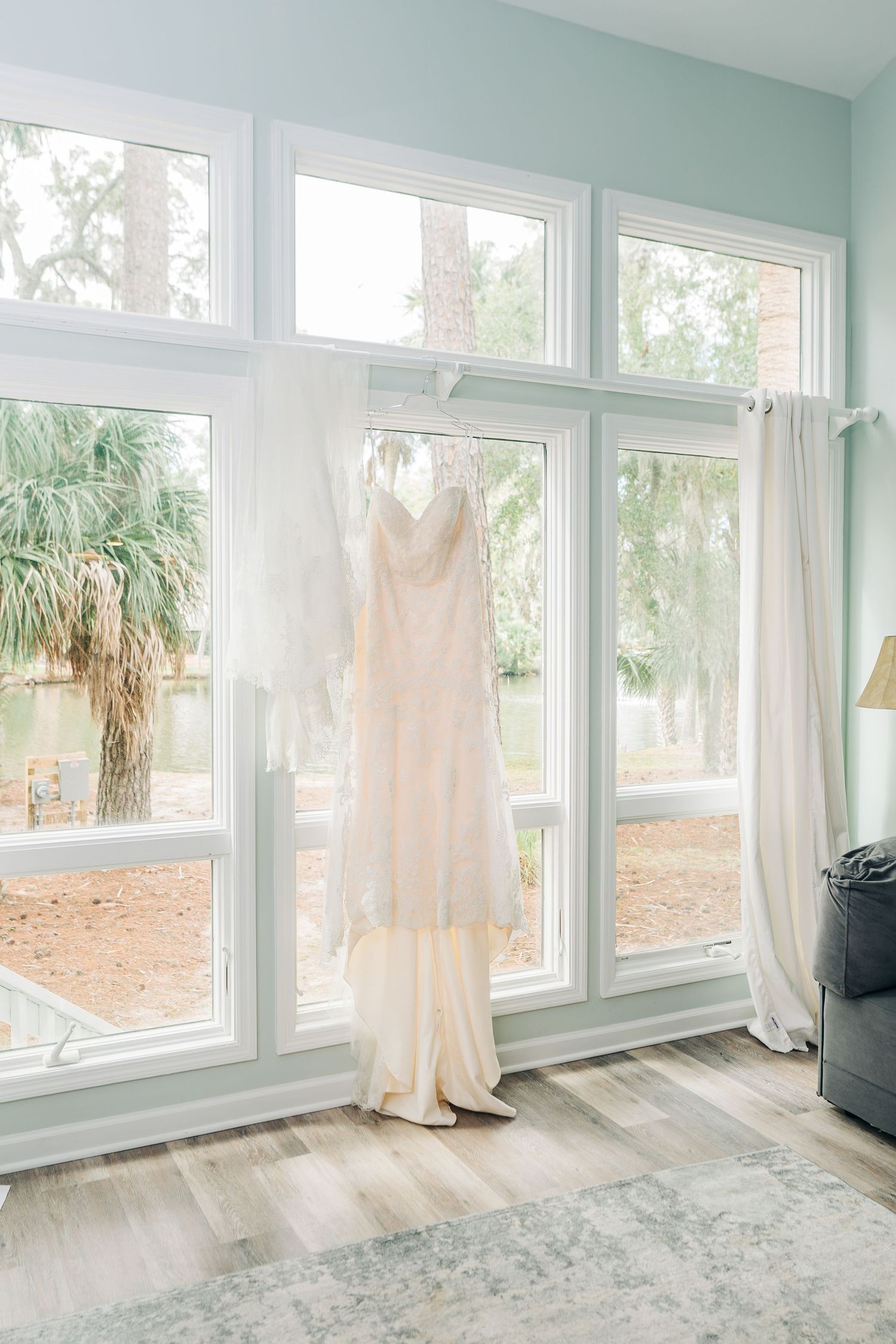 An elegant white wedding dress hangs in front of bright windows in a modern room with mint green walls.