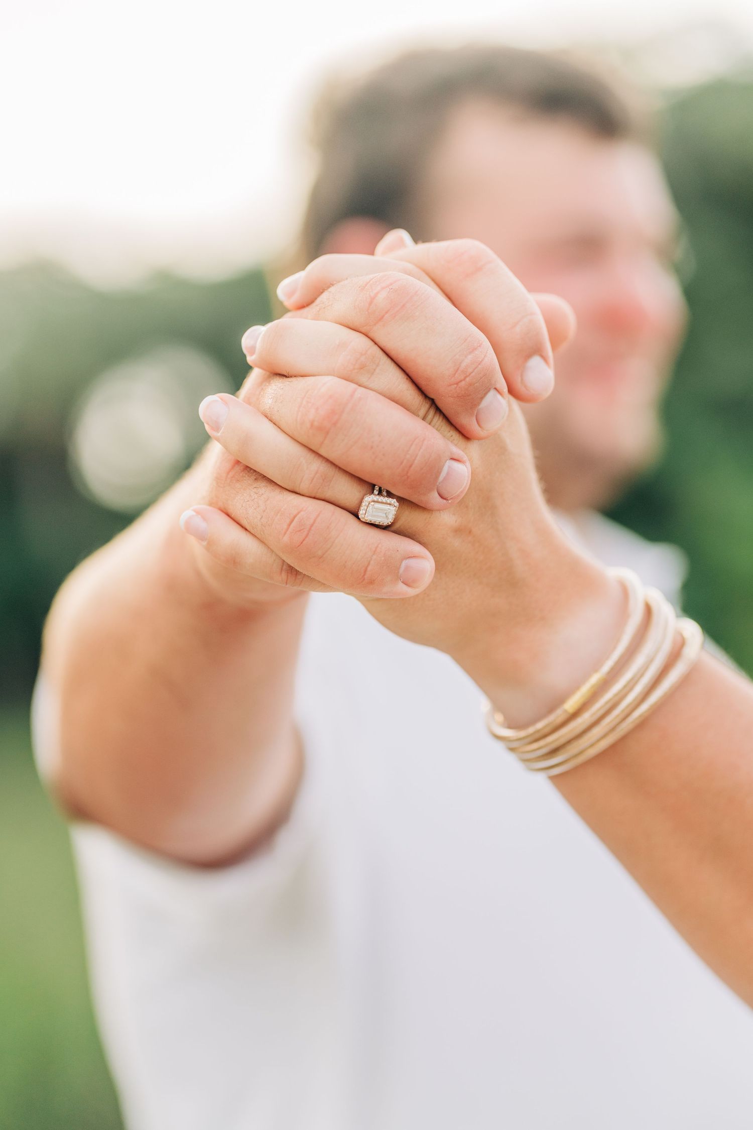 Close-up detail of hands with diamond engagement ring and delicate gold bangles against soft outdoor background.