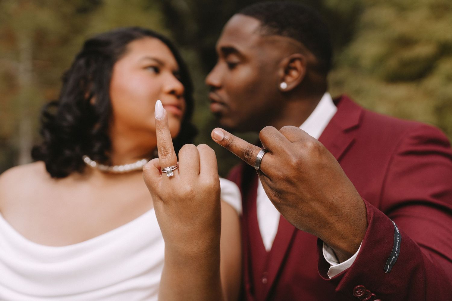A couple in burgundy and white formal attire share a tender moment during an outdoor photoshoot.