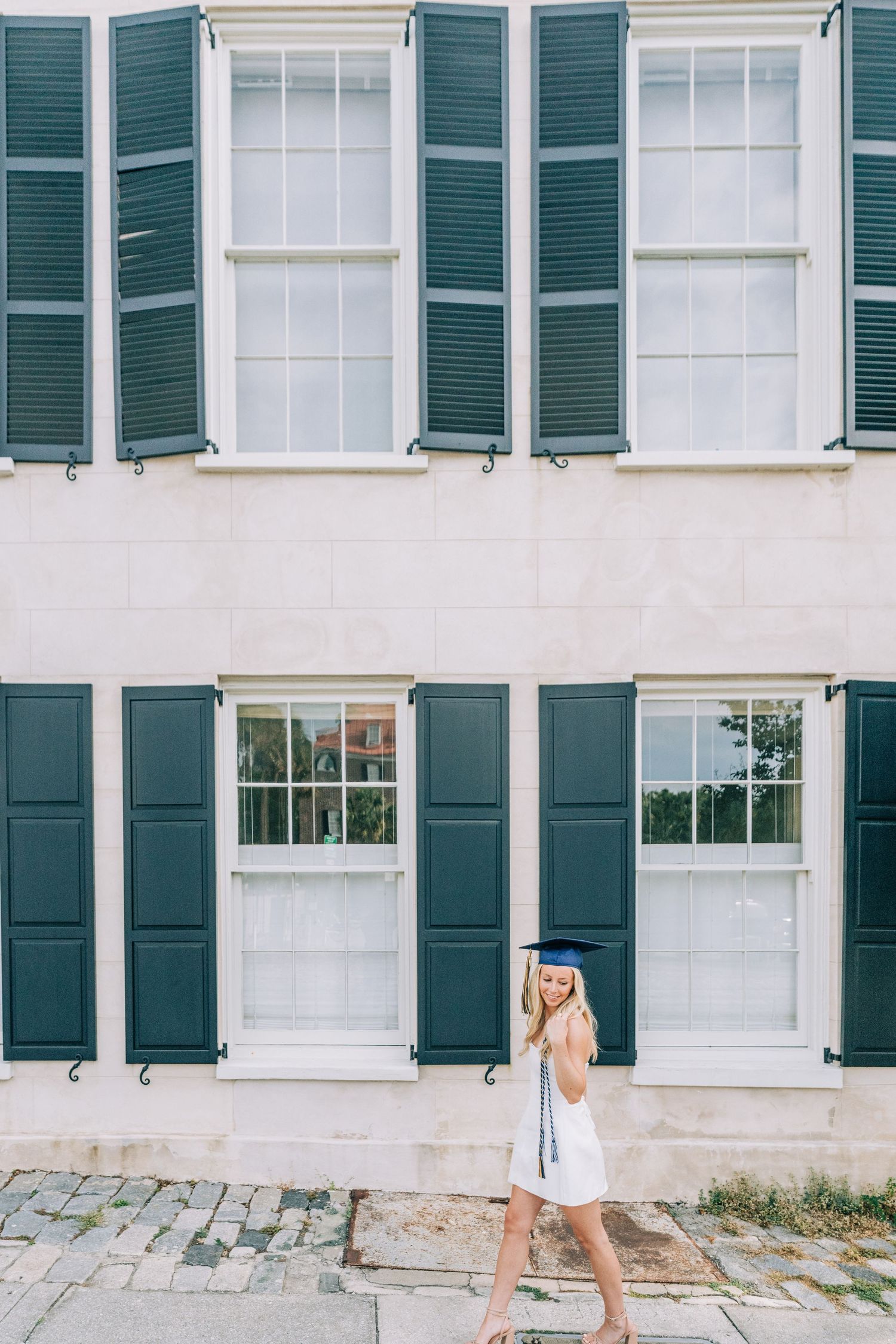 Woman in white dress walks past historic building with green shuttered windows.