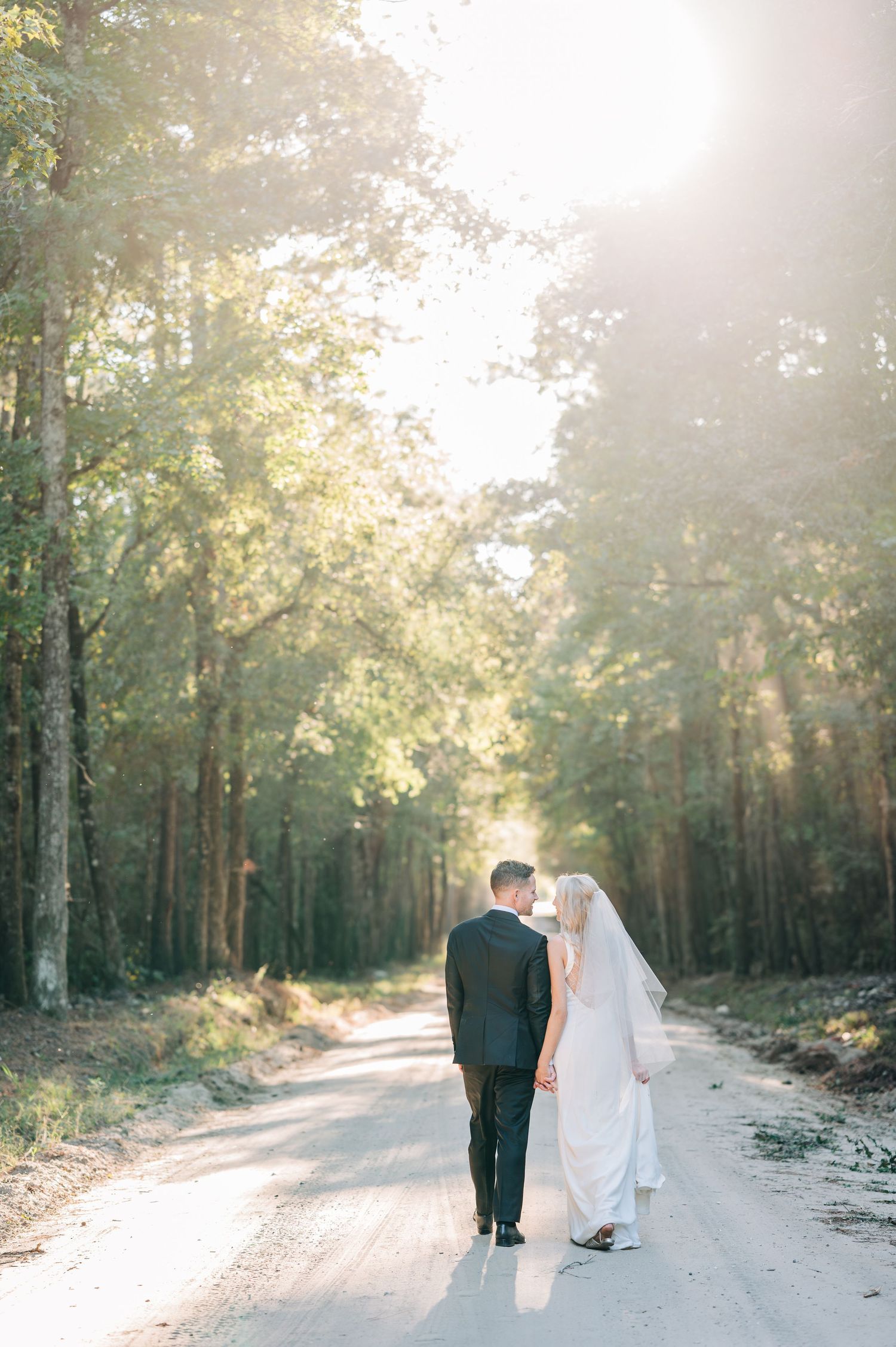 Couple walking down sunlit forest path in wedding attire.