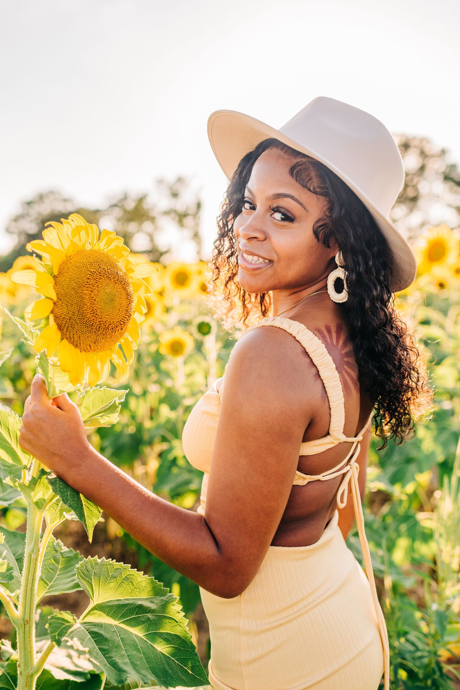Woman in white hat poses playfully with bright yellow sunflowers in a sunny field.