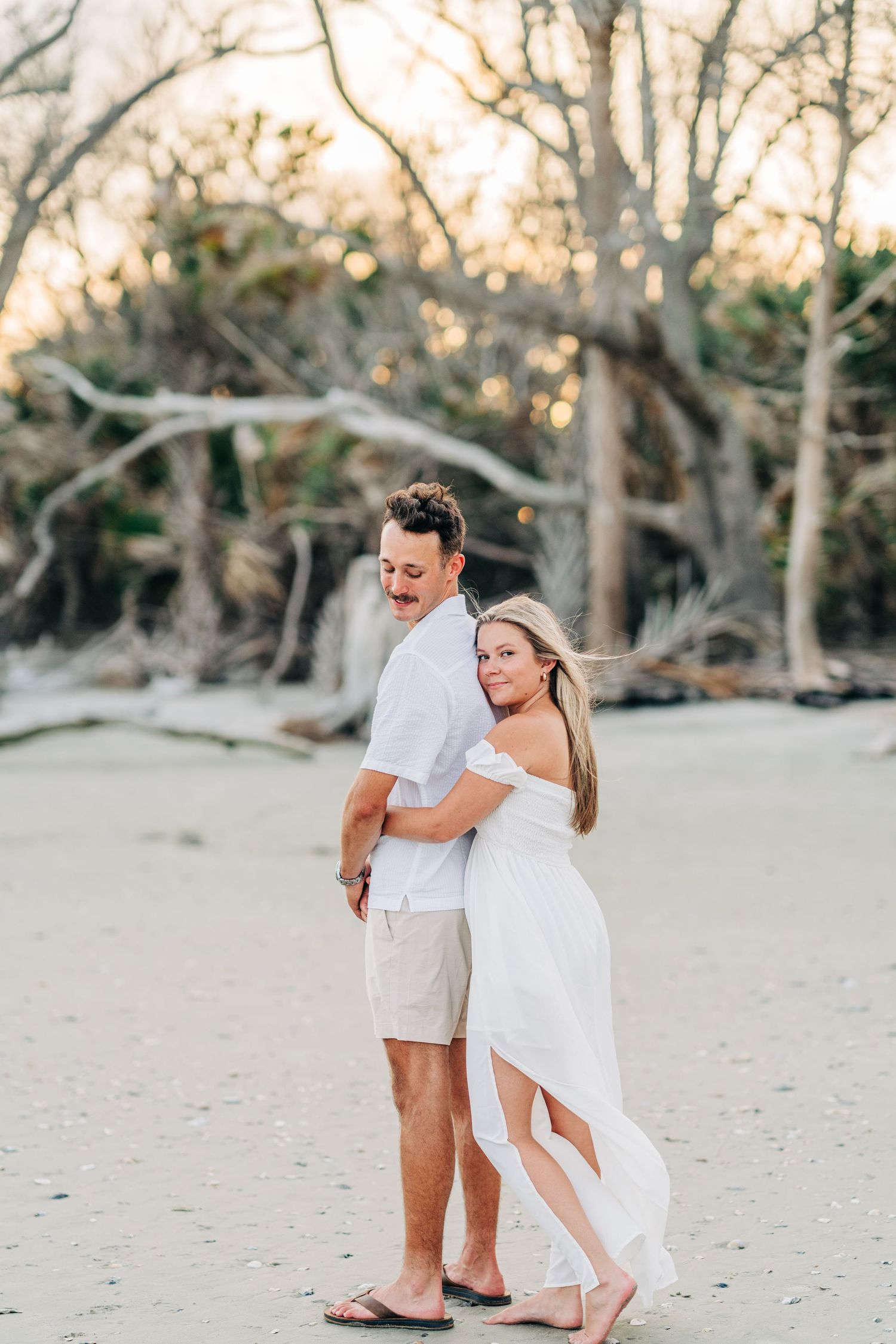 A romantic couple walking together on a sandy beach at sunset wearing white coastal attire.