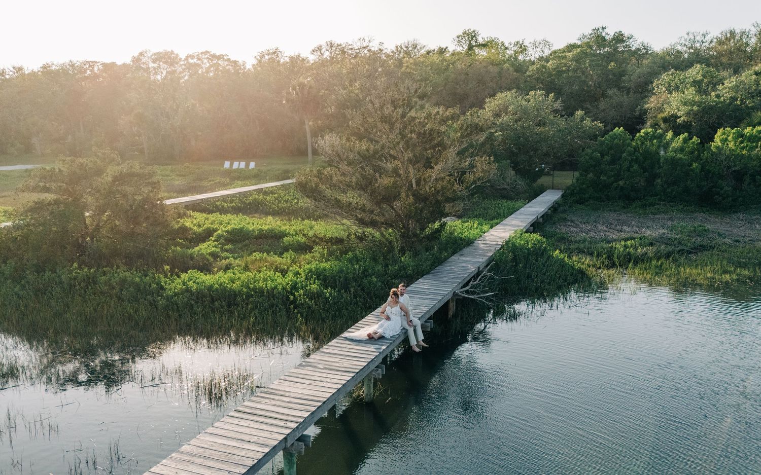 Wooden boardwalk stretches across a marshy waterway during golden sunset hour.