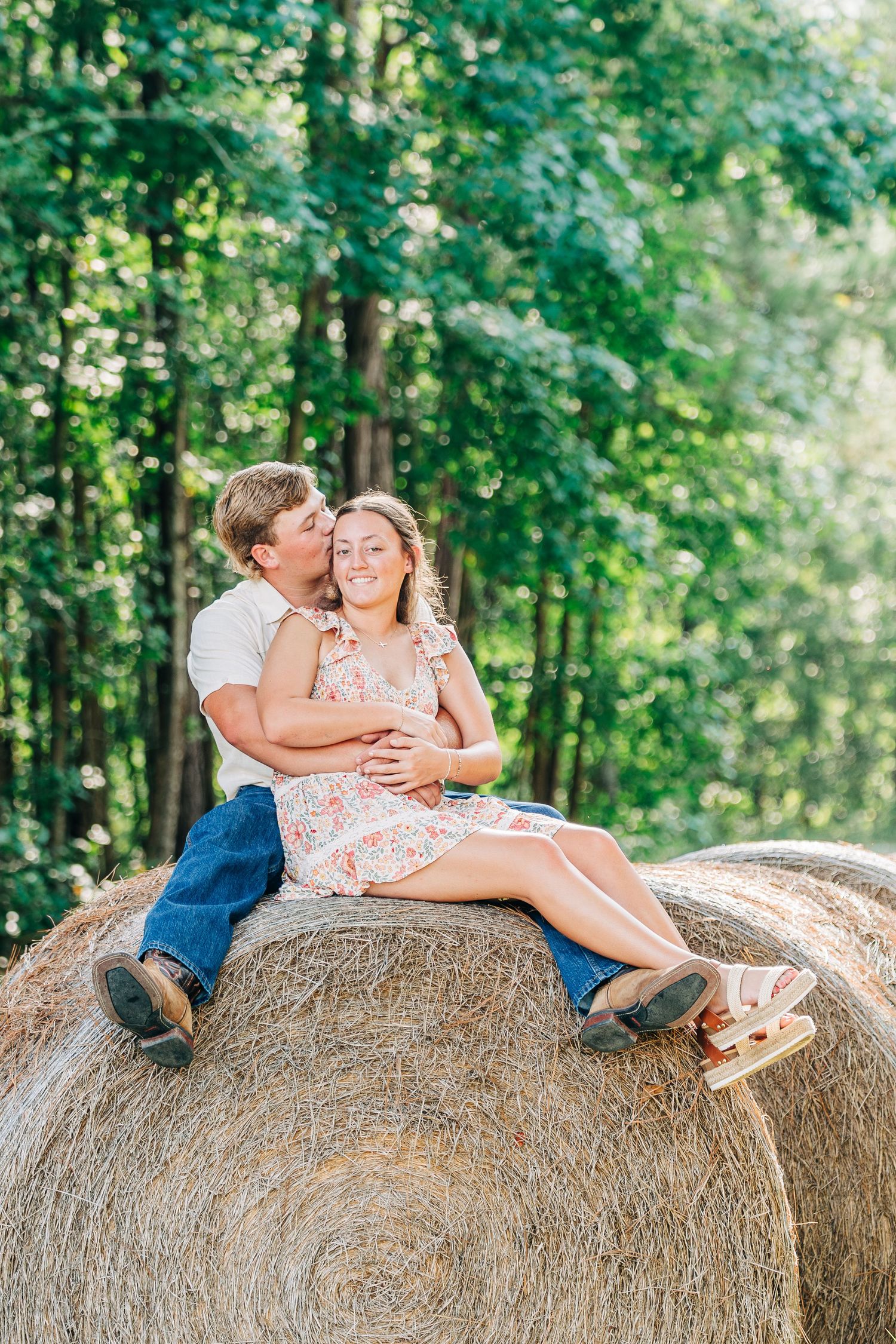 Couple enjoying romantic moments together while sitting on large round hay bales in a sunny field.