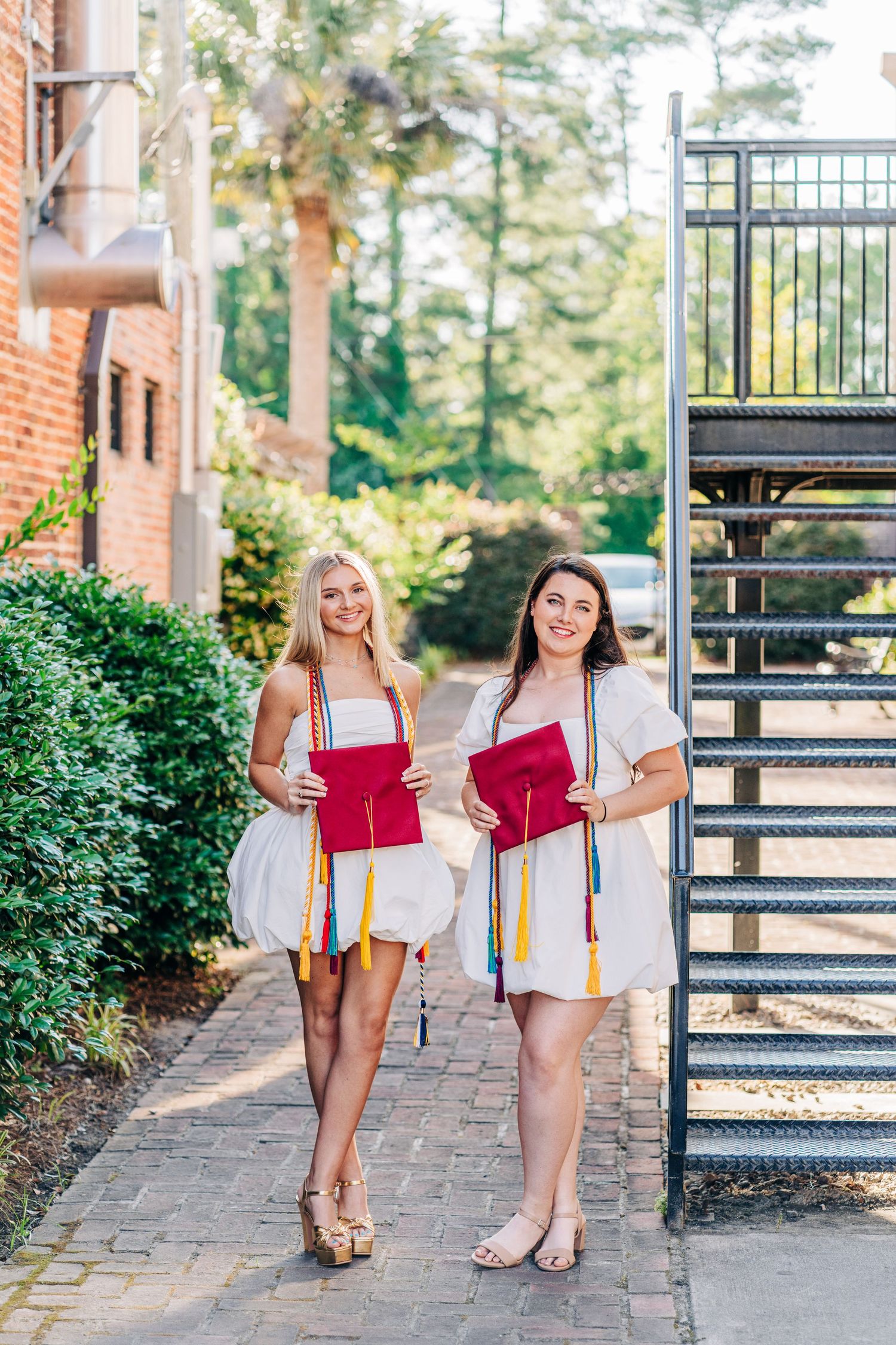 Two graduates in white dresses hold red diploma covers while standing on a brick pathway.