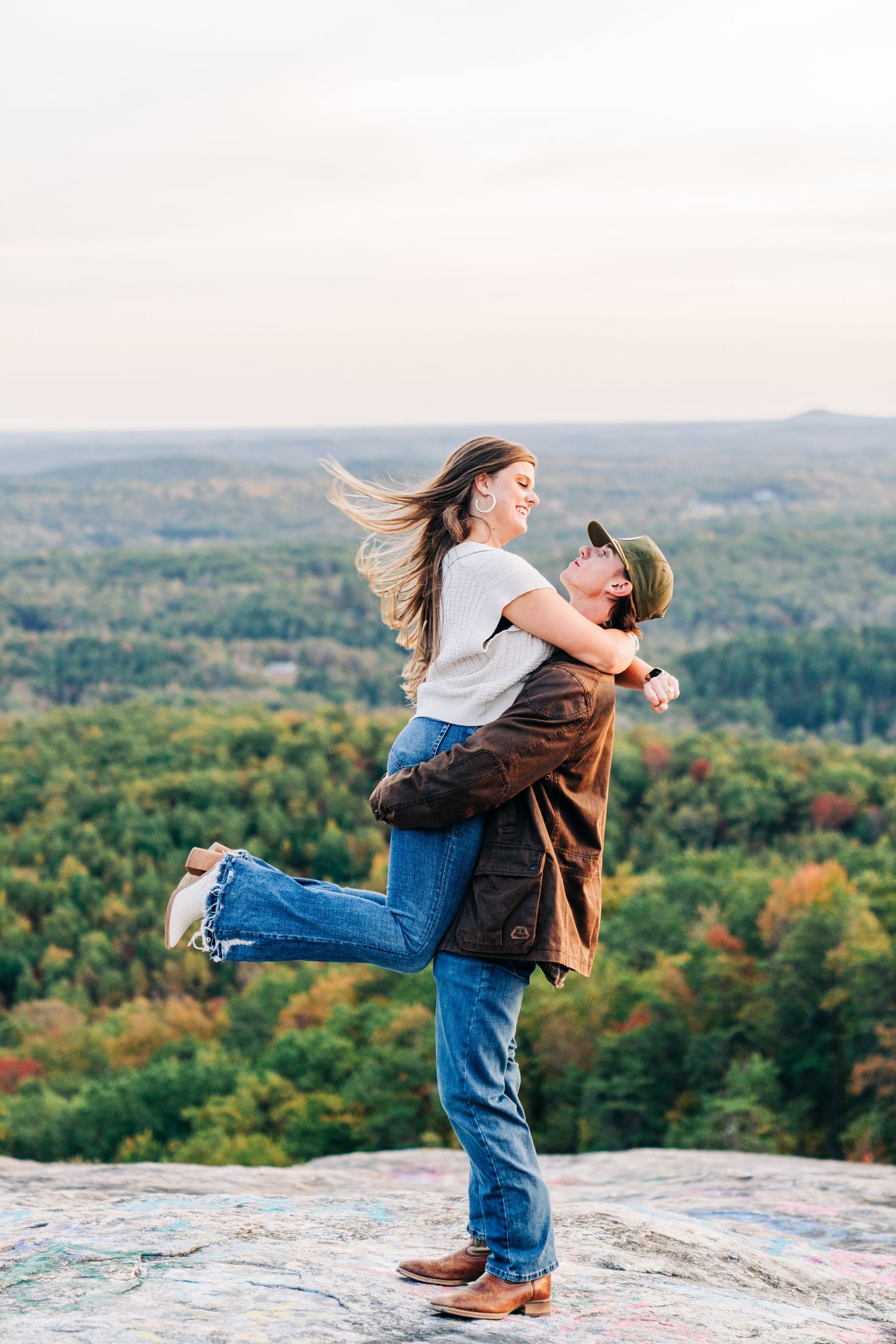 Couple embraces and spins joyfully on a scenic mountain overlook during autumn with forest views in background.