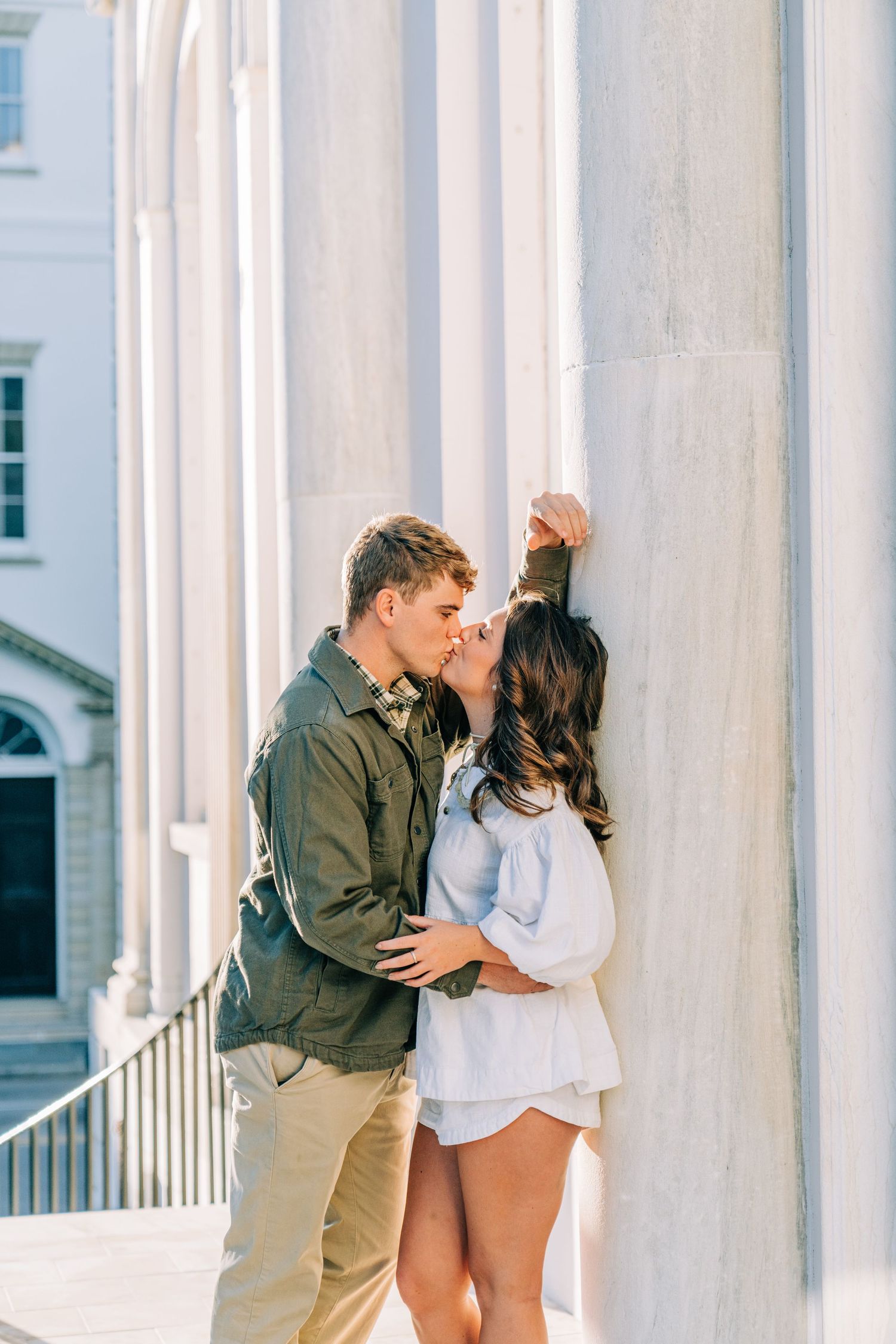 Romantic couple embracing between white columns in elegant architectural setting during engagement photoshoot.