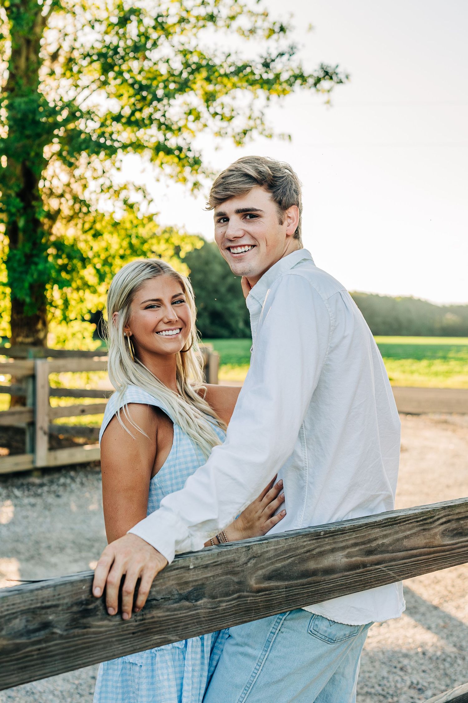 Couple embracing by a wooden fence at golden hour in a rustic countryside setting.