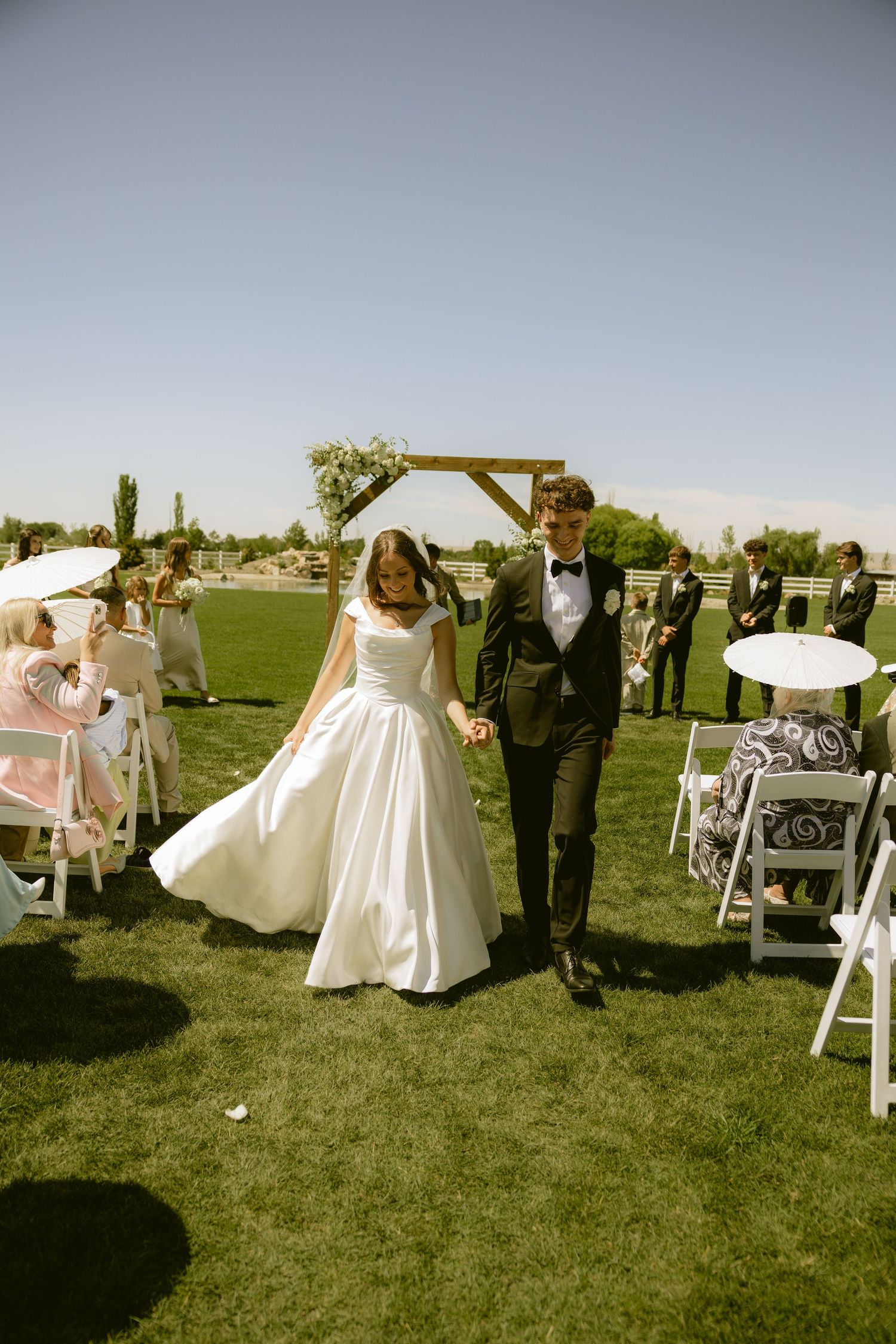 Bride and groom walk hand-in-hand down outdoor wedding aisle on green lawn with wooden arch backdrop.