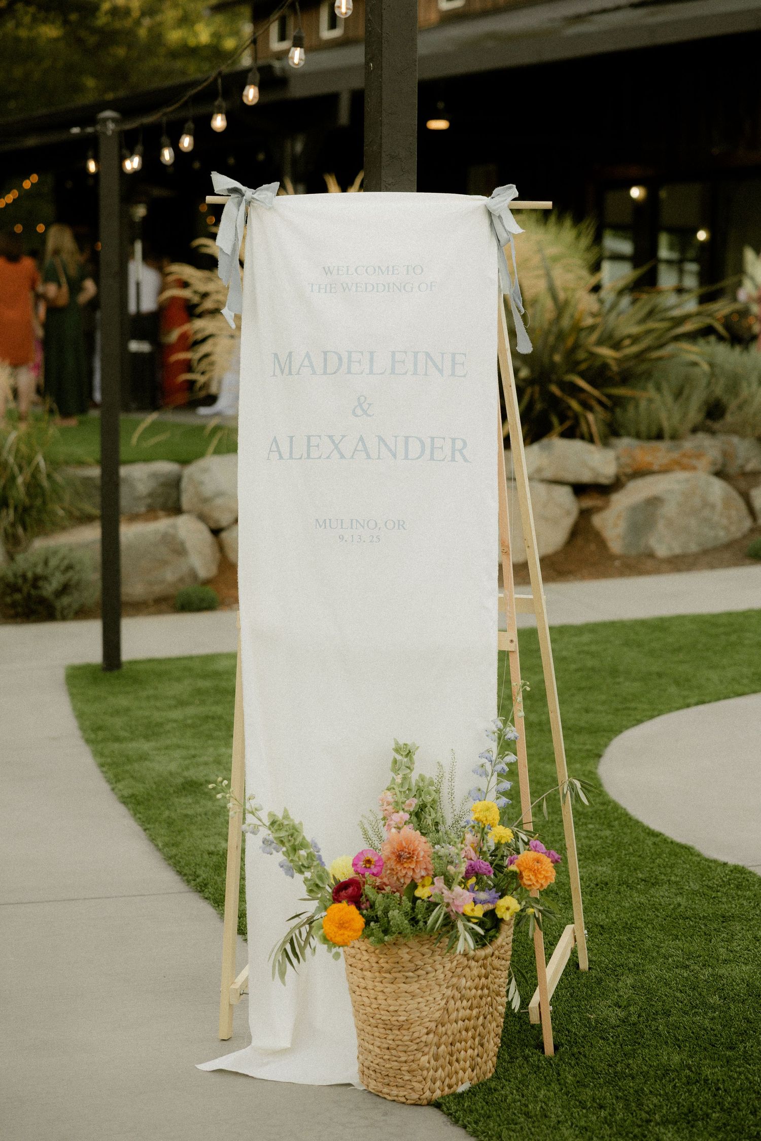 Wedding welcome sign with floral decorations and woven basket on garden pathway.