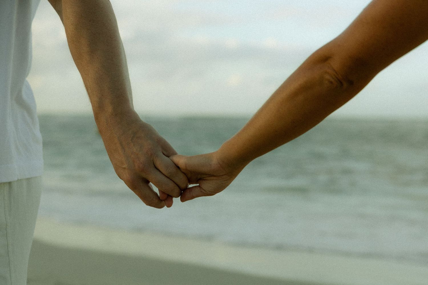 Two people hold hands tenderly on a peaceful beach with ocean waves in the background.