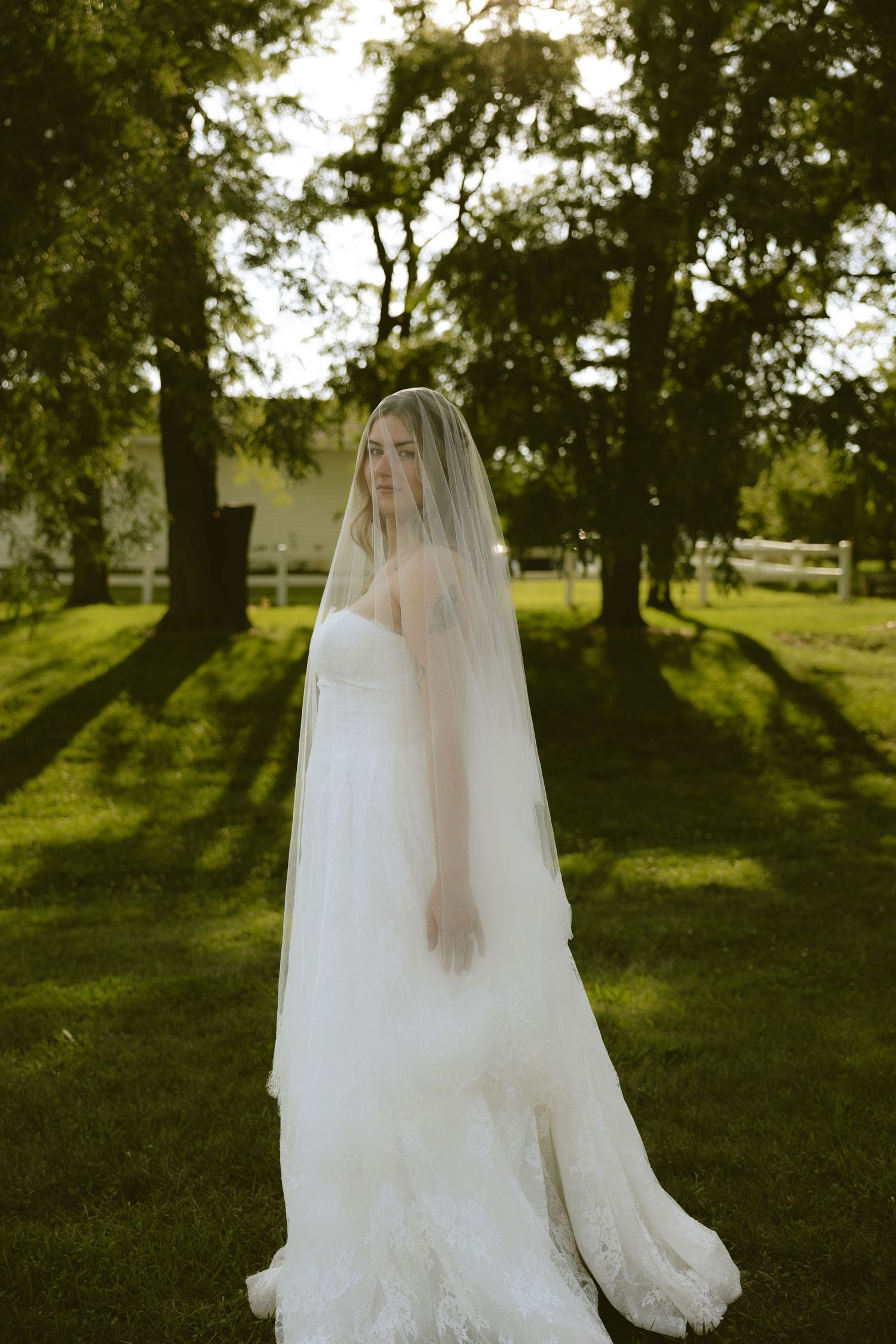 Bride in strapless lace gown and flowing cathedral veil stands on lush green lawn surrounded by trees.