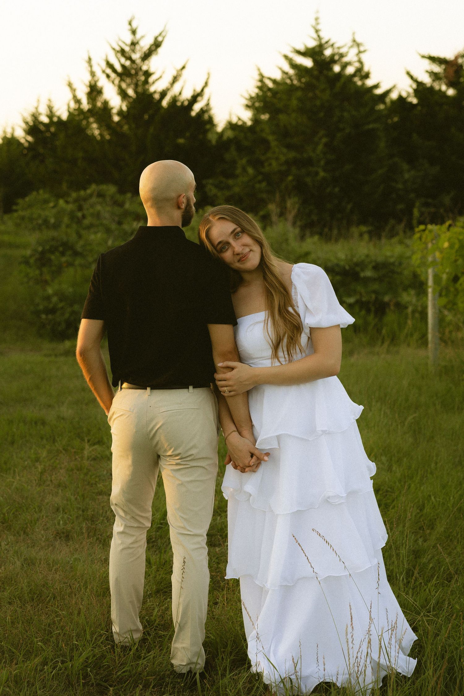 Bride in white tiered dress leans on groom's shoulder while holding hands in a lush green field at sunset.