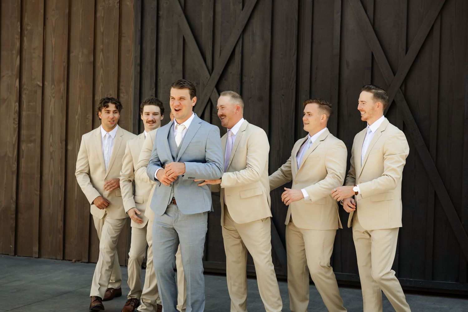 Groomsmen in light gray suits share candid moments against dark backdrop.