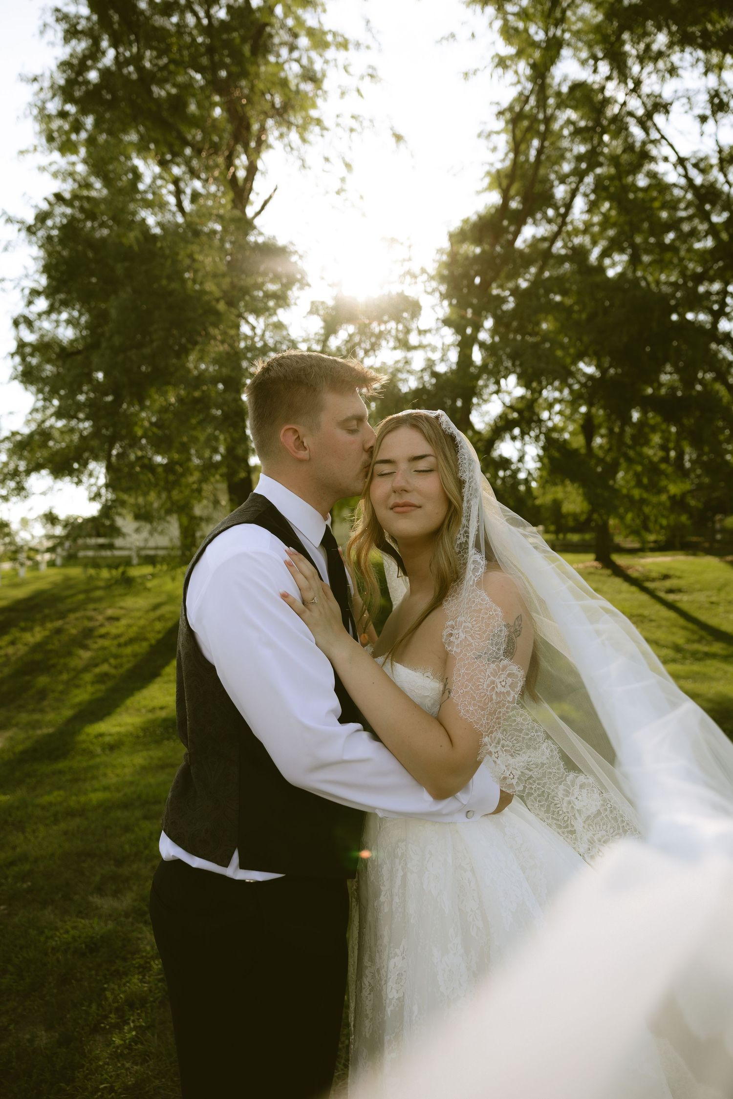 Groom kissing bride in golden sunlight outdoors, bride in lace gown with veil, surrounded by lush green trees.