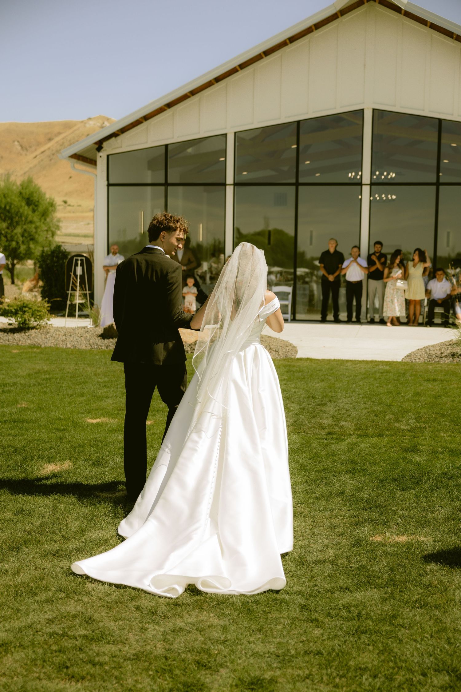 Bride and groom stand together outdoors during wedding ceremony on green lawn in front of modern white barn venue.
