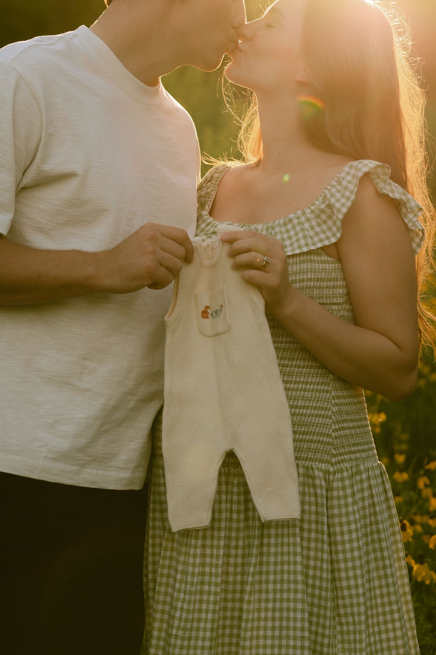 Couple kissing at golden hour holding tiny baby overalls, announcing pregnancy outdoors in sunflower field.