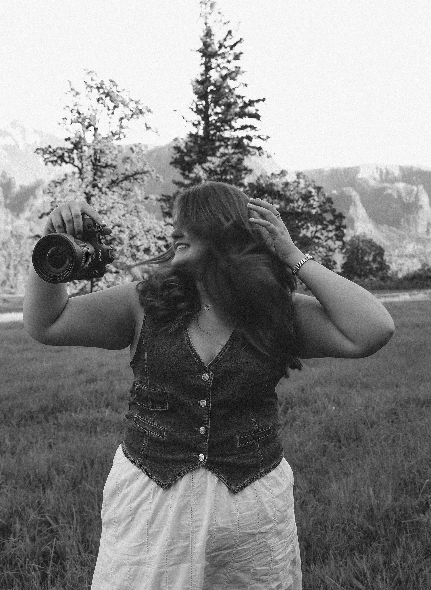 Black and white photo of a woman holding a camera outdoors, wearing a denim vest, with trees and mountains behind her.