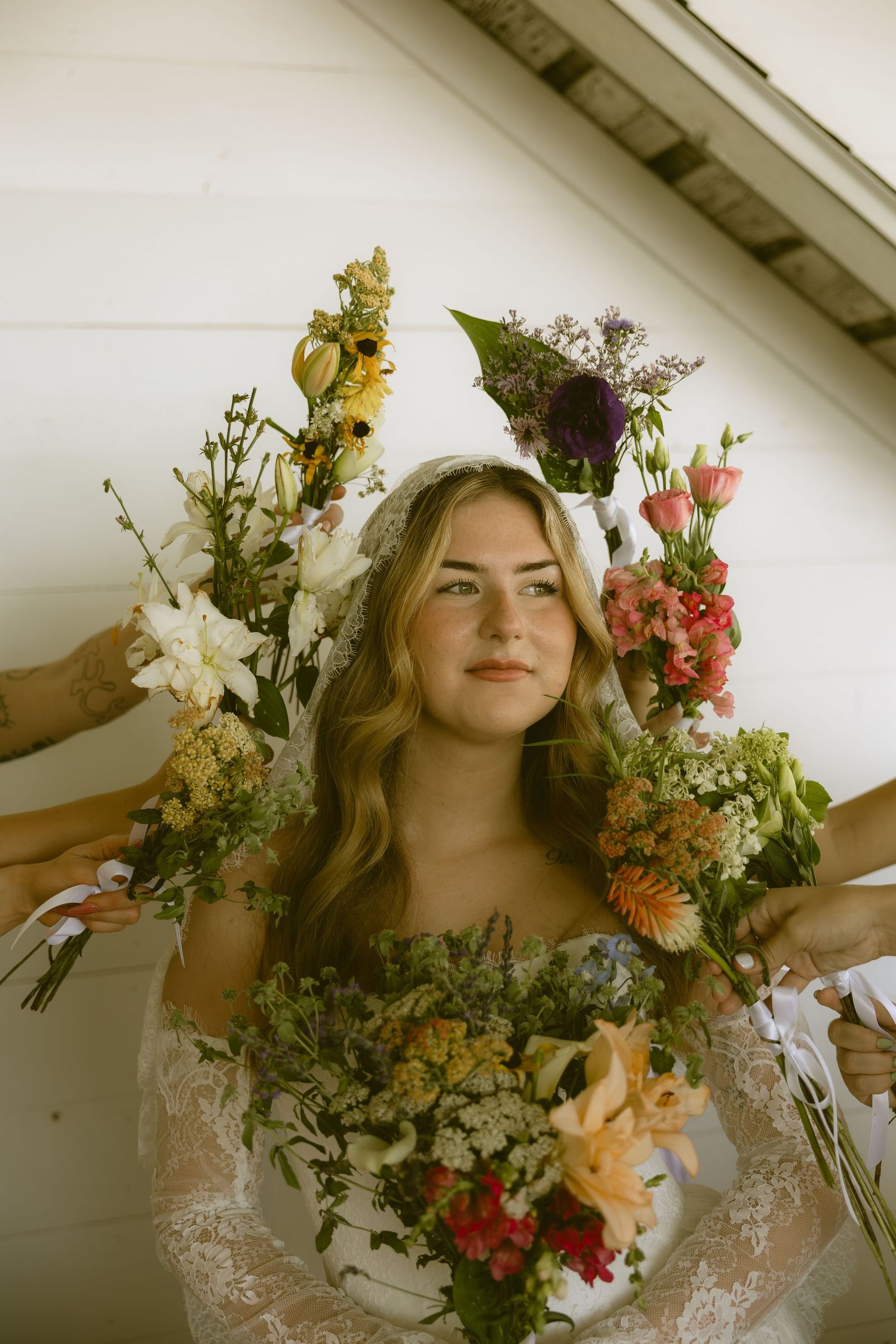Bride in a lace gown surrounded by colorful wildflower bouquets held by bridesmaids against a white wood backdrop.