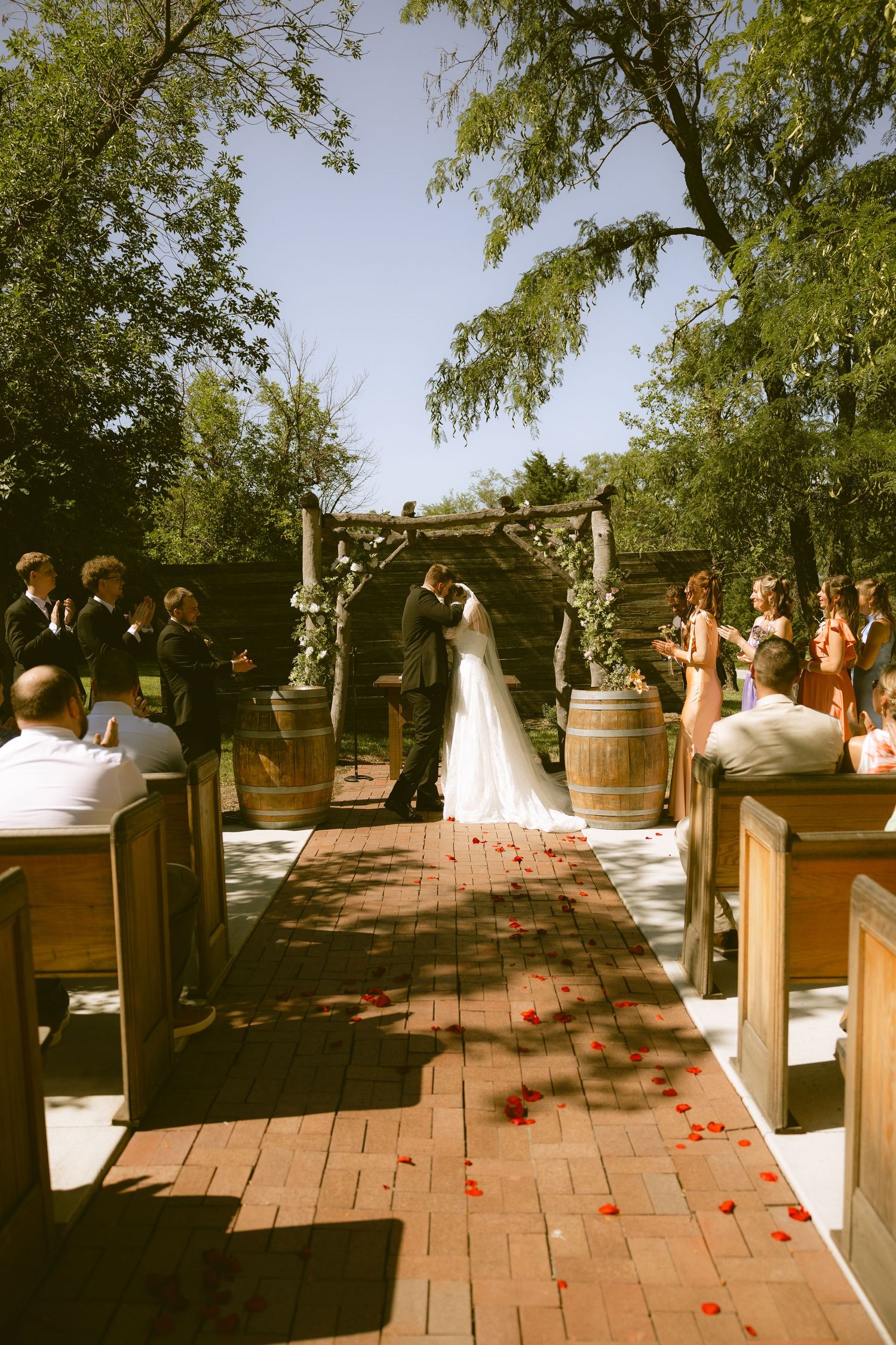Outdoor wedding ceremony on brick aisle with rose petals, couple kissing at rustic wooden arch altar surrounded by guests.