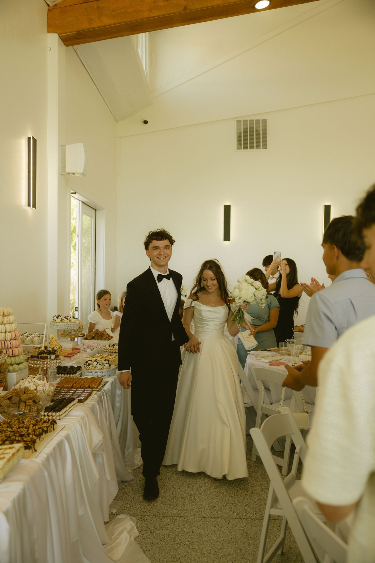 Smiling newlyweds make grand entrance into bright white wedding reception hall past dessert table.
