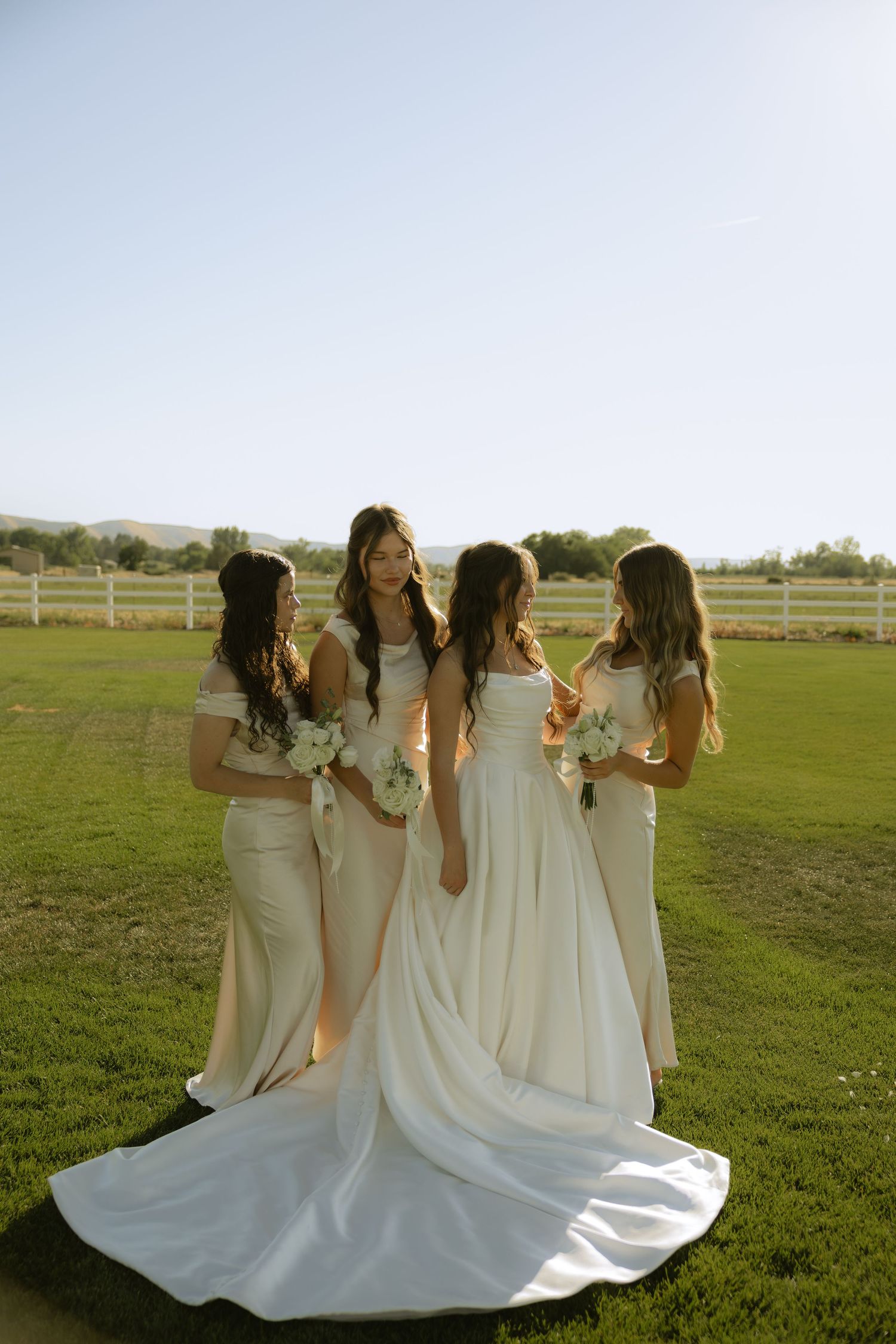 Bride and three bridesmaids in ivory gowns holding white bouquets pose on green farm lawn.