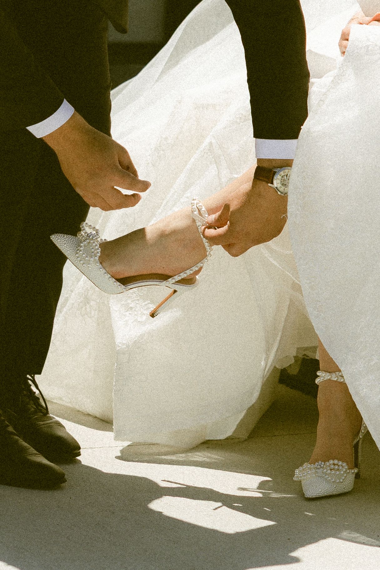 Groom helping bride put on pearl-embellished white wedding heels, with white bridal gown and black dress shoes visible.