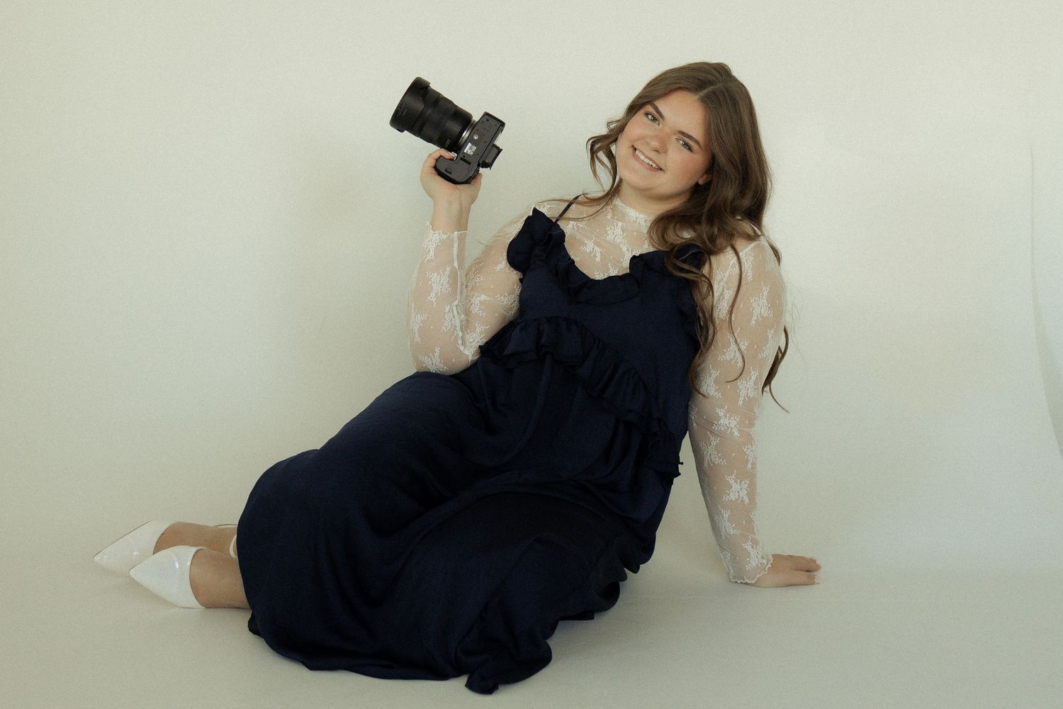A photographer in a dark dress reclines while holding a camera in a bright studio setting.
