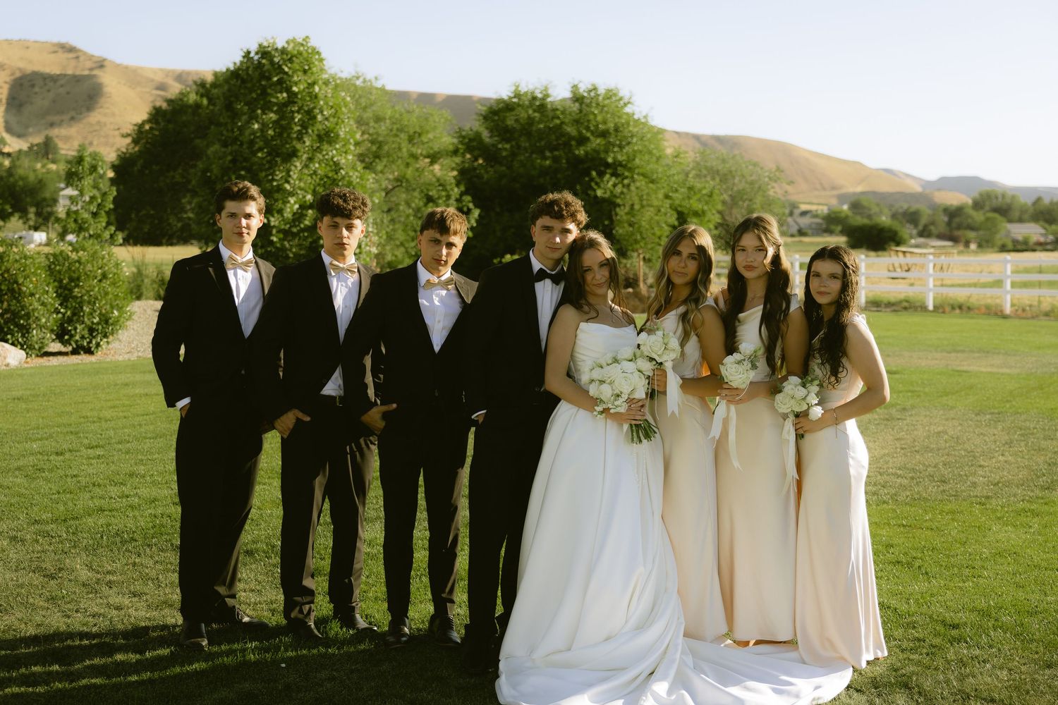 Wedding party poses outdoors with bride in white gown, groom, groomsmen in black suits, and bridesmaids in ivory dresses.