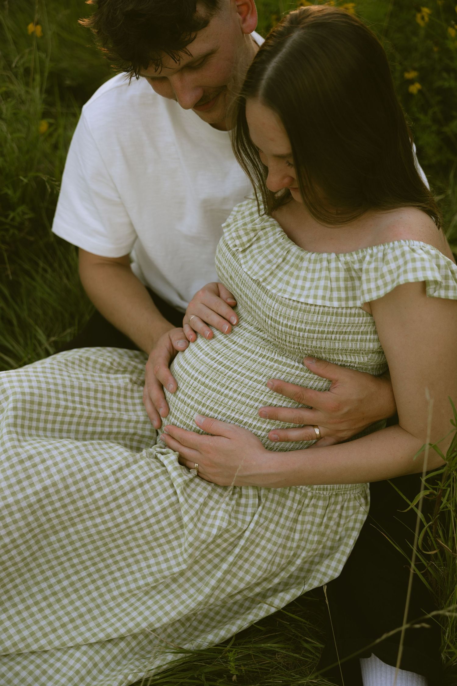 Couple sitting in green field, man embracing pregnant woman in green gingham off-shoulder dress during golden hour.