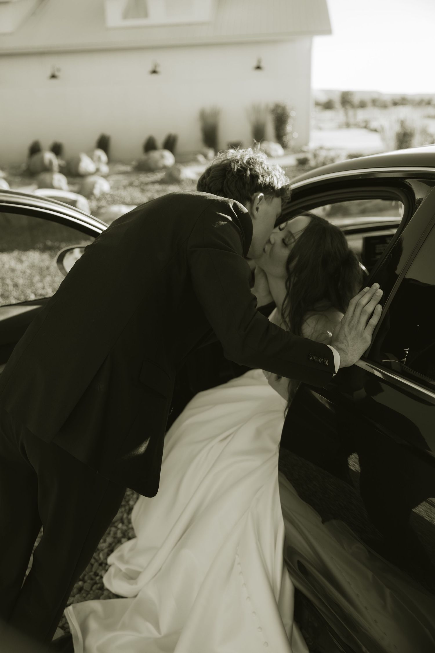 Groom kissing bride in white dress through car window in black and white photo.