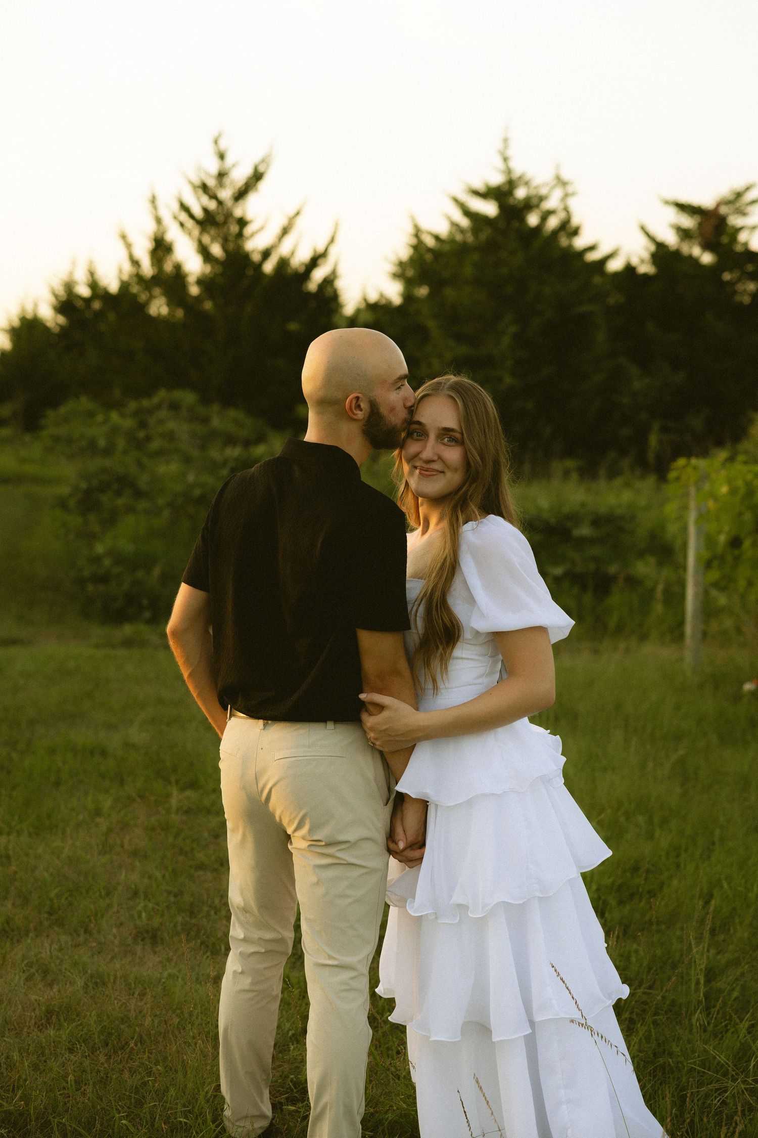 Groom in black shirt kissing bride on cheek, she smiles in white tiered dress in a green field at golden hour.