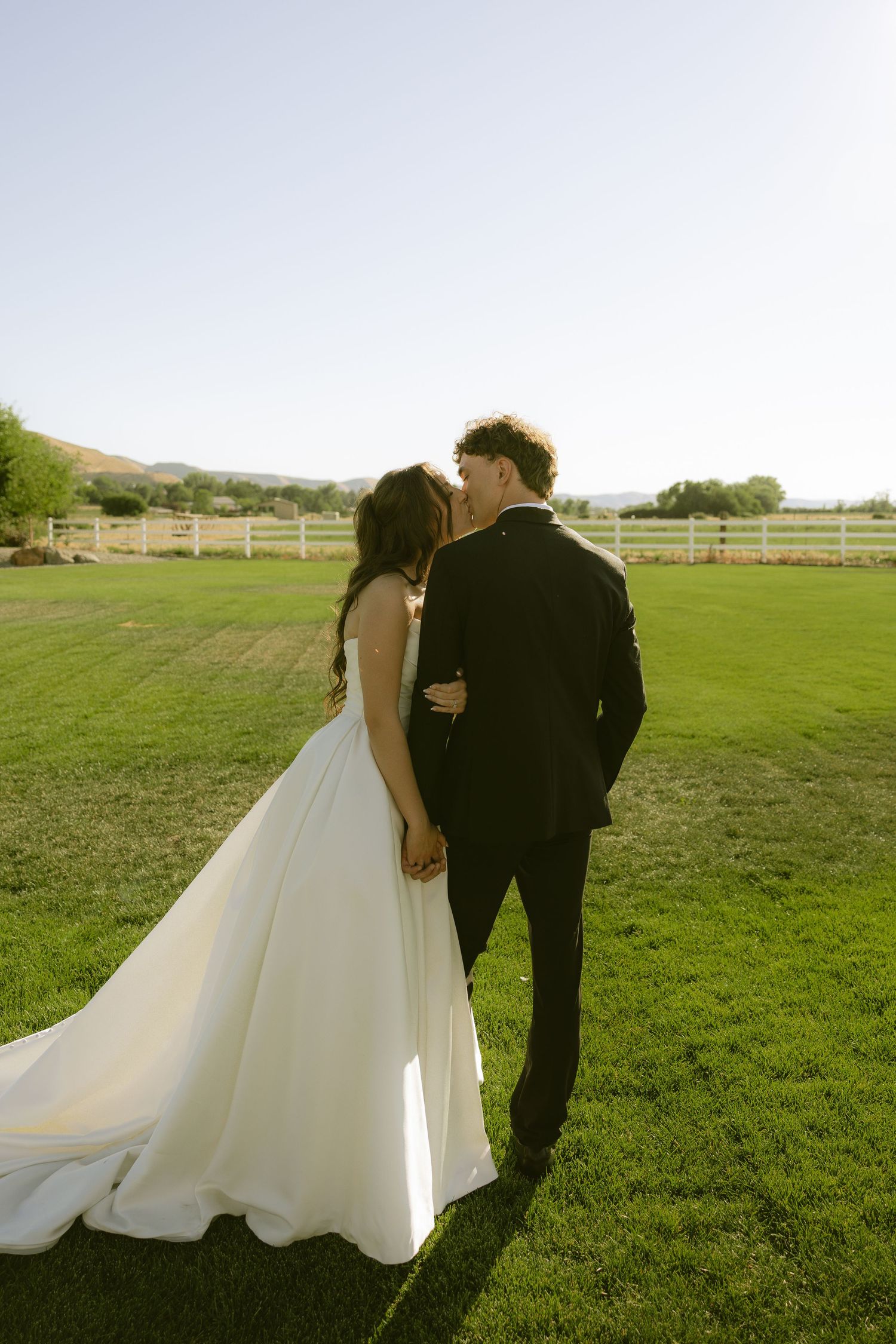 A bride and groom share a kiss outdoors on a green lawn with a white fence and rolling hills in the background.