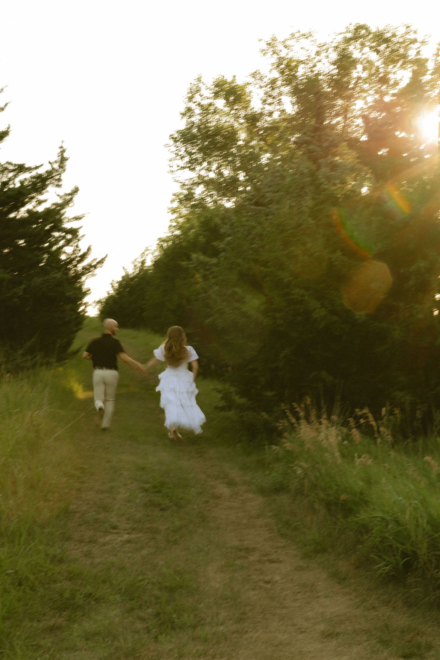 Bride and groom holding hands running down a sunlit garden path surrounded by lush green trees at golden hour.