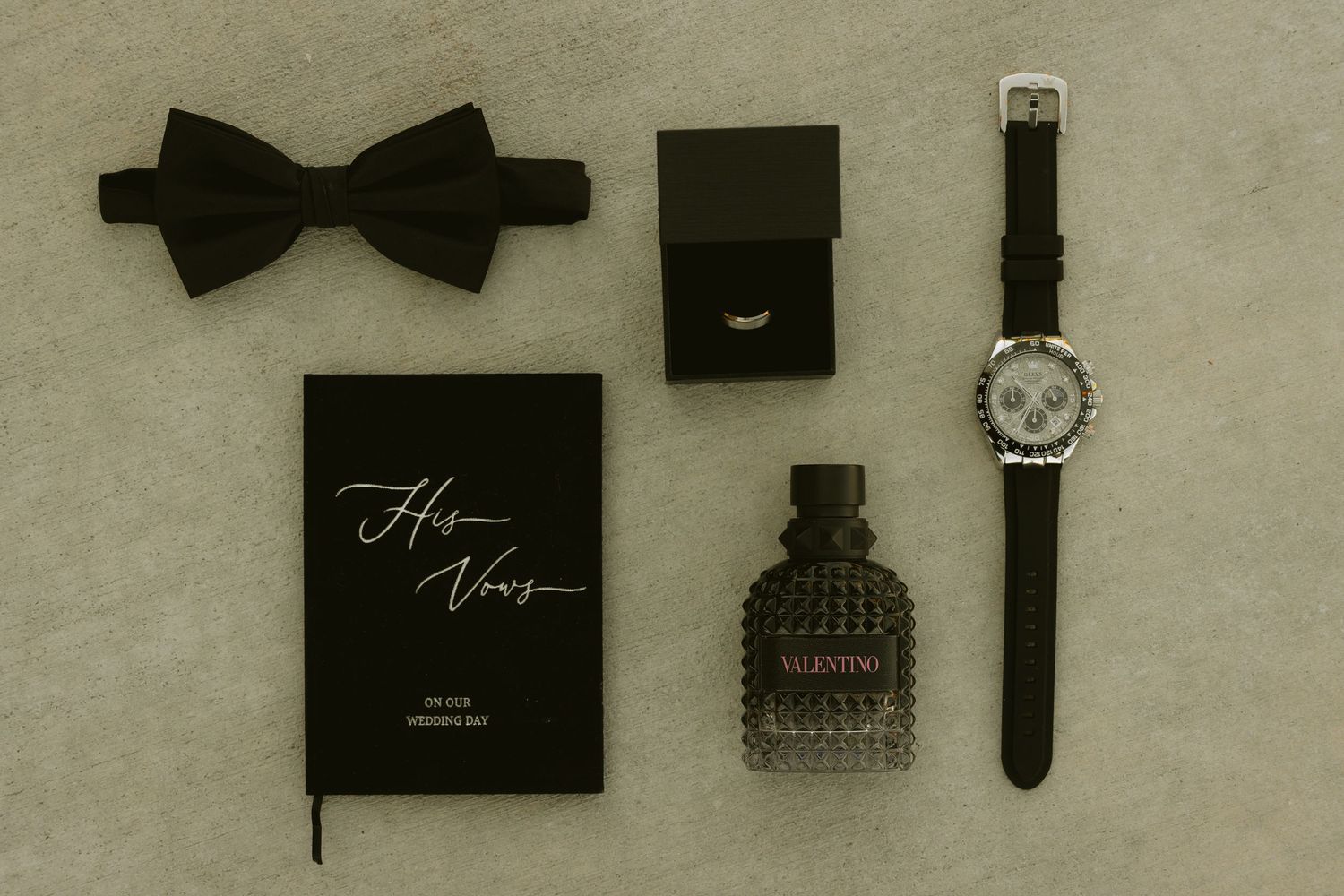 Groom flat lay featuring black bow tie, watch, ring box, cologne, and vow book.