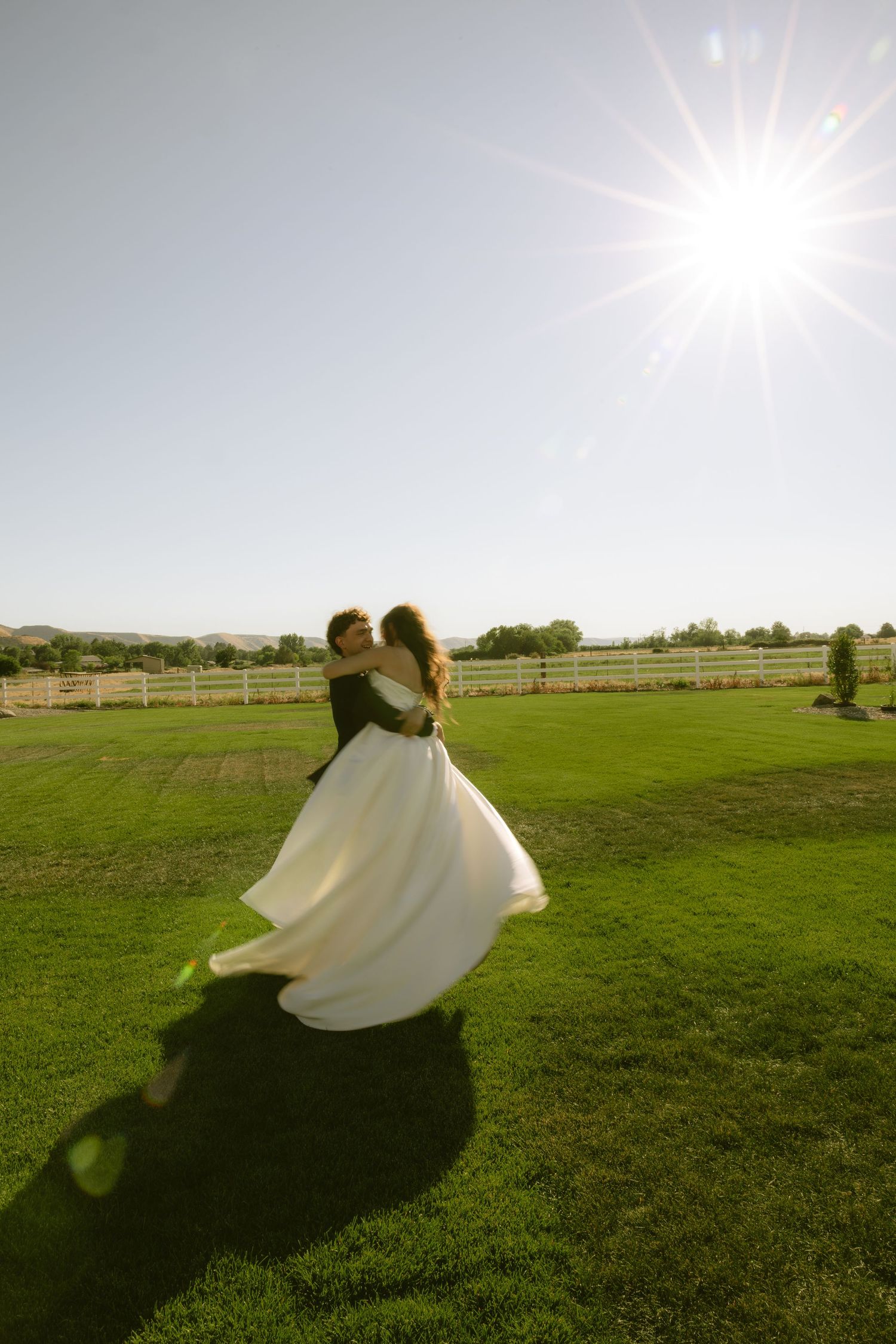 Groom lifting bride in white gown on sunny green lawn at outdoor wedding venue.